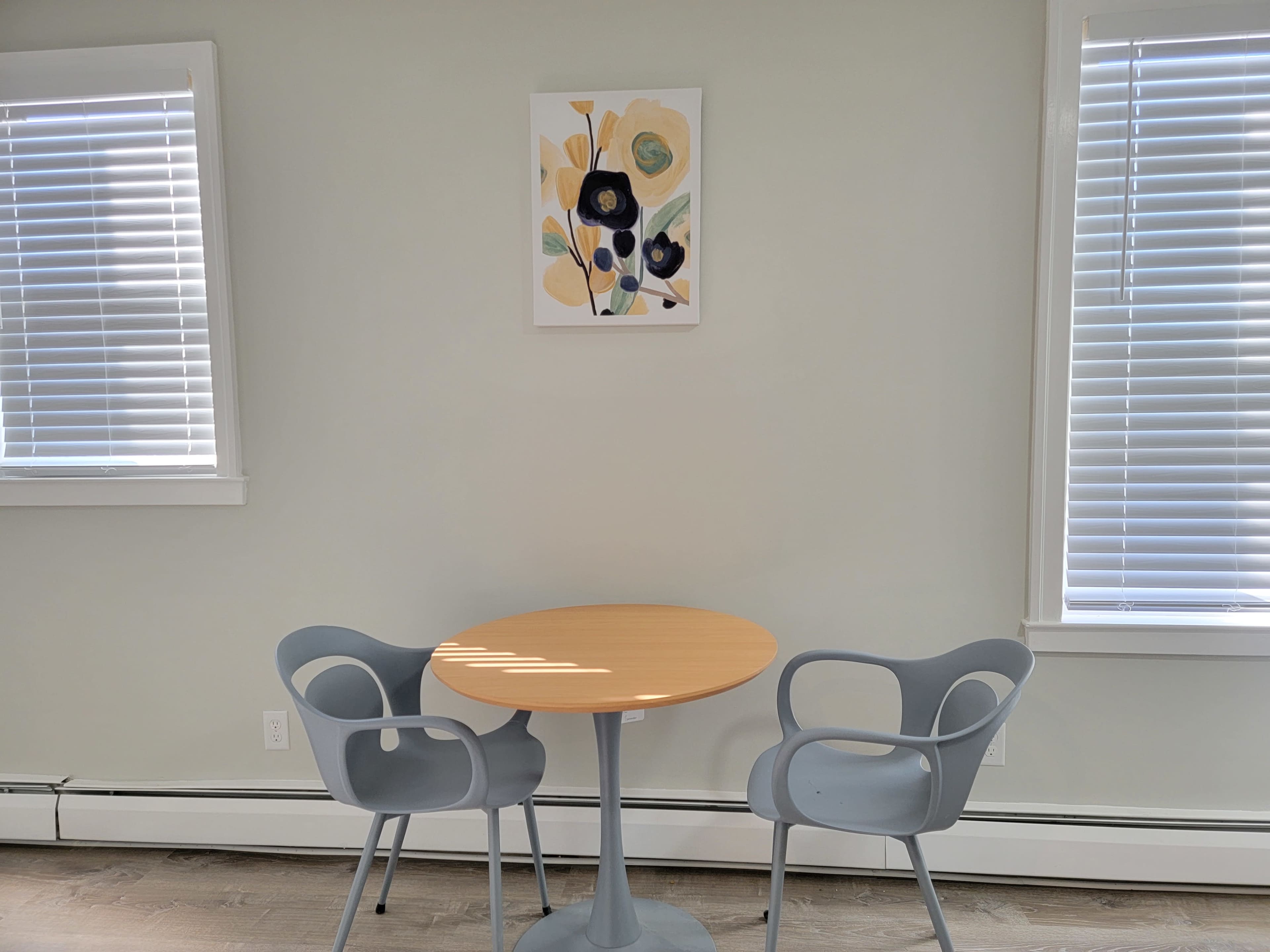 A small round table with two chairs is positioned against a light gray wall, adorned with a floral artwork and flanked by two windows with blinds.