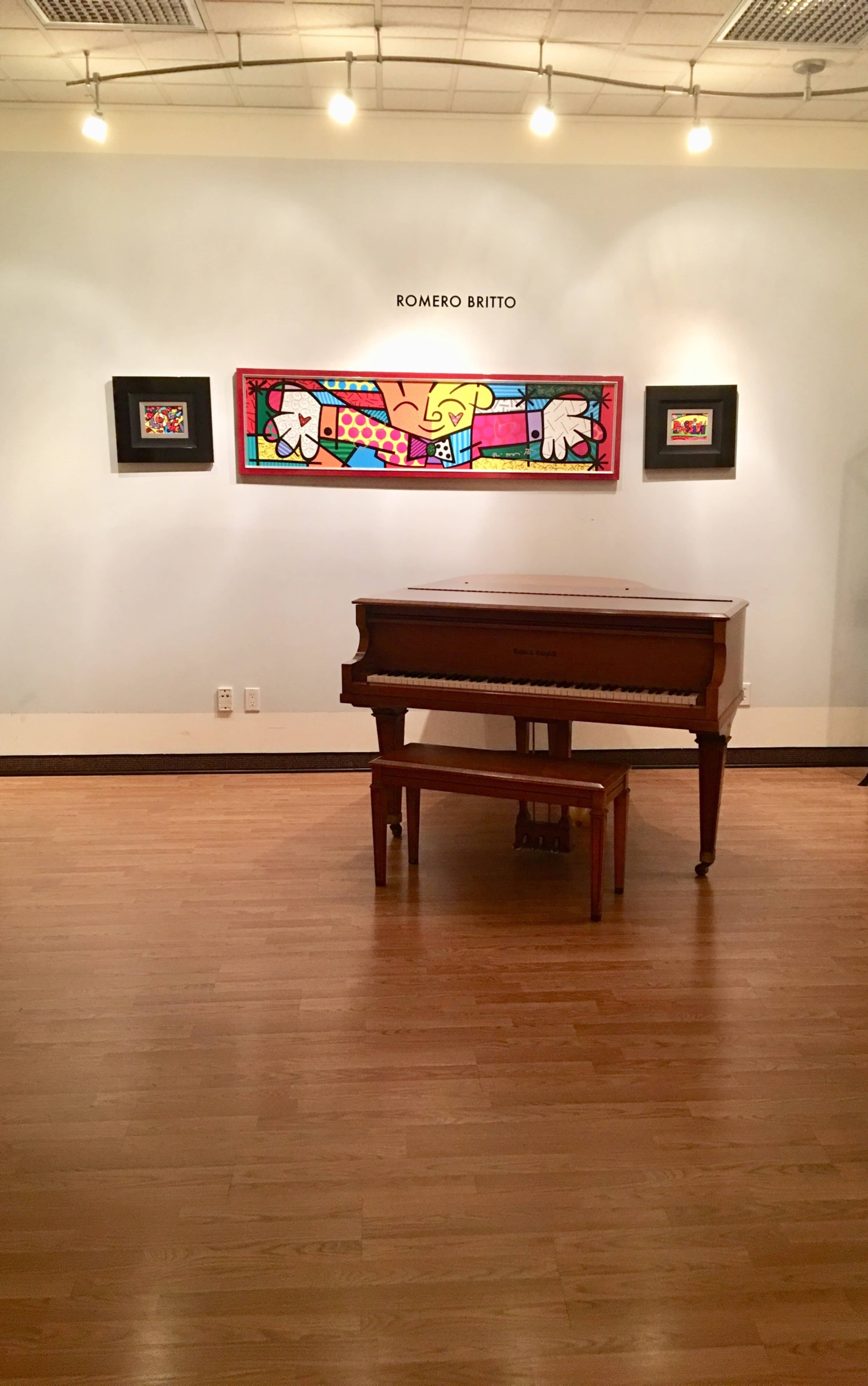 A wooden piano is situated in a gallery beneath a colorful artwork by Romero Britto on the wall.
