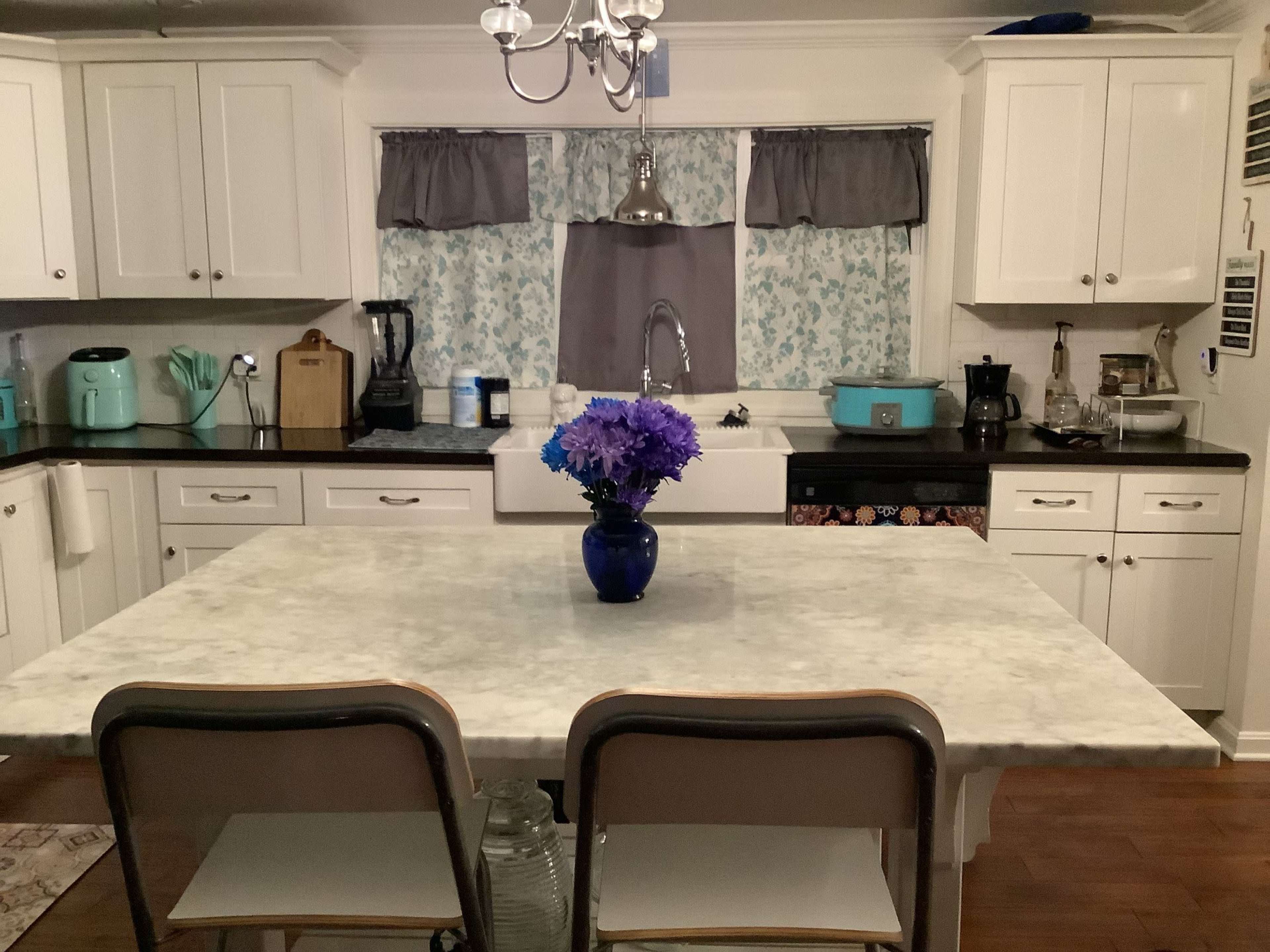 A kitchen features a marble island with two chairs, a vase of purple flowers, and various appliances against white cabinetry.