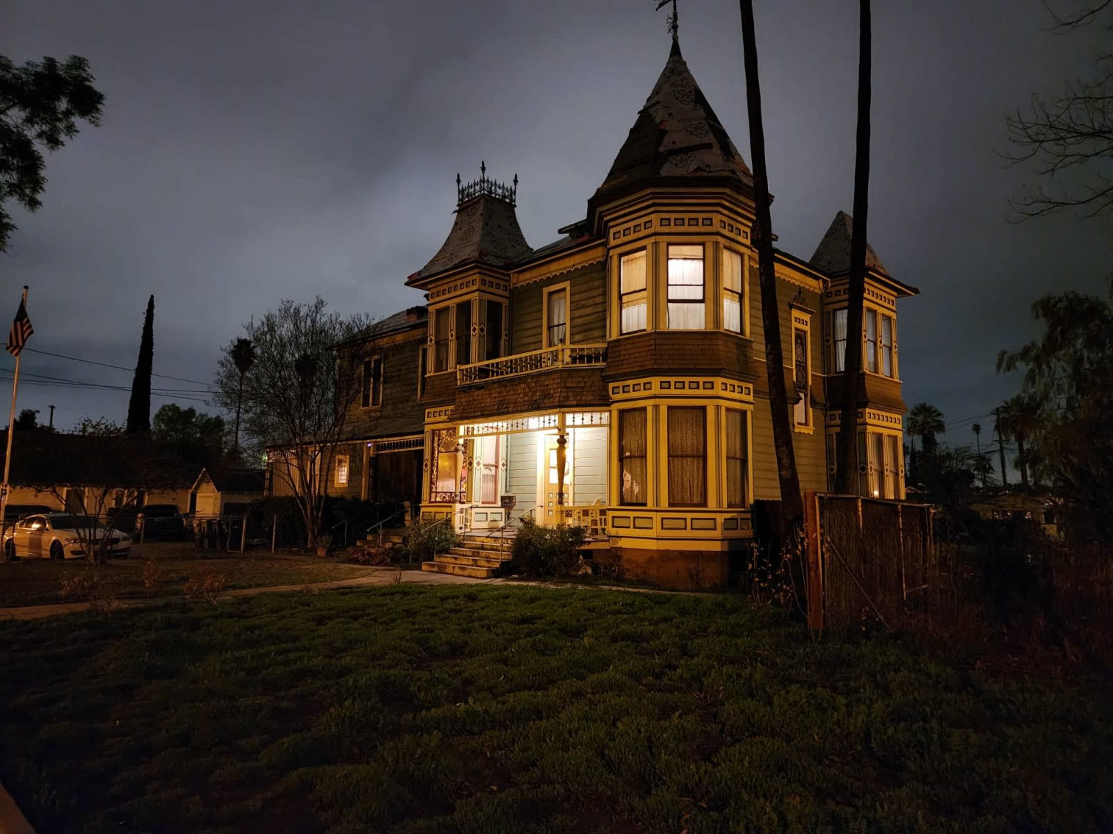 A large, two-story Victorian house with a pointed turret is illuminated at night, surrounded by sparse greenery and trees.