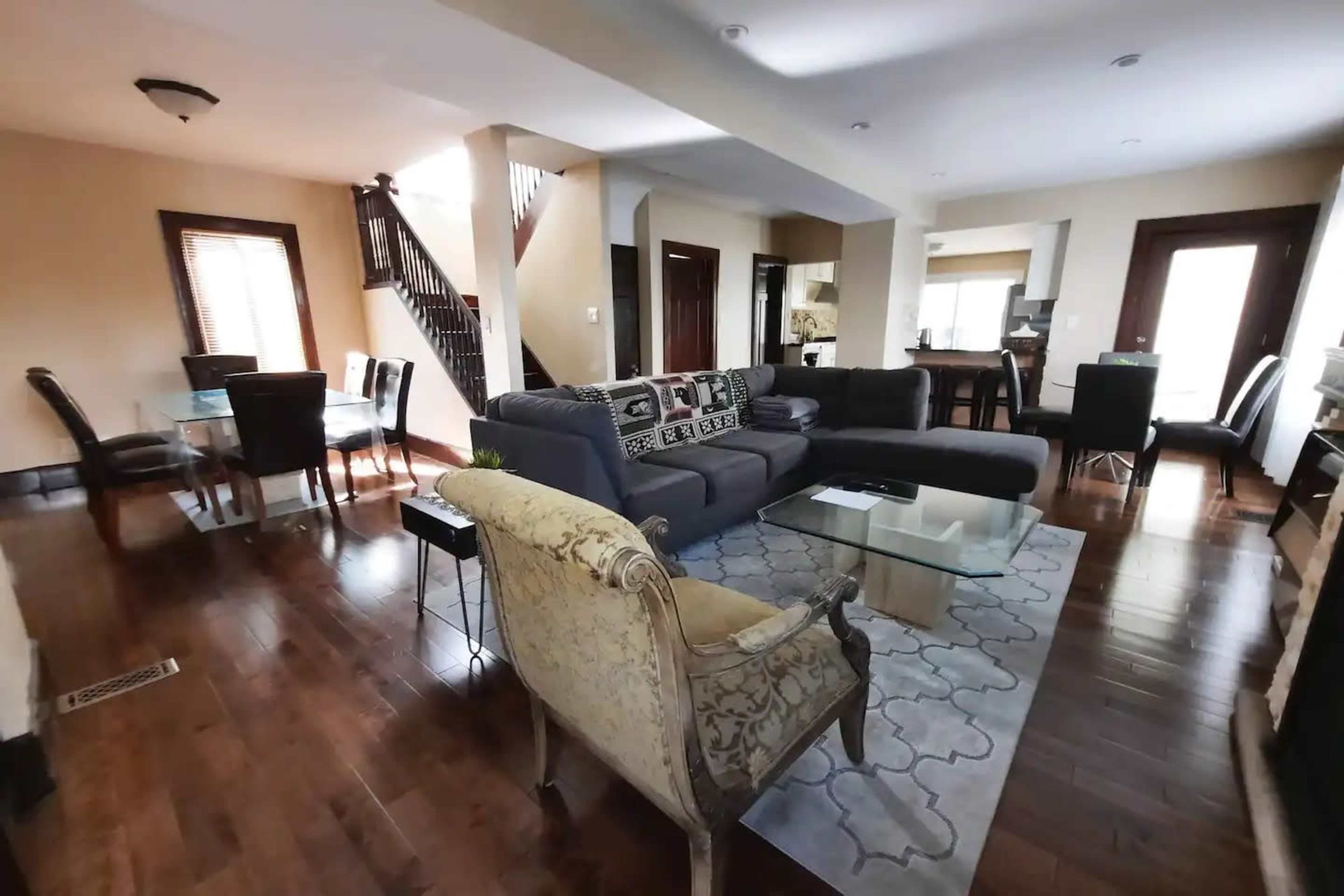 A spacious living area with a sectional sofa, a glass coffee table, a dining table, and a staircase visible in the background.