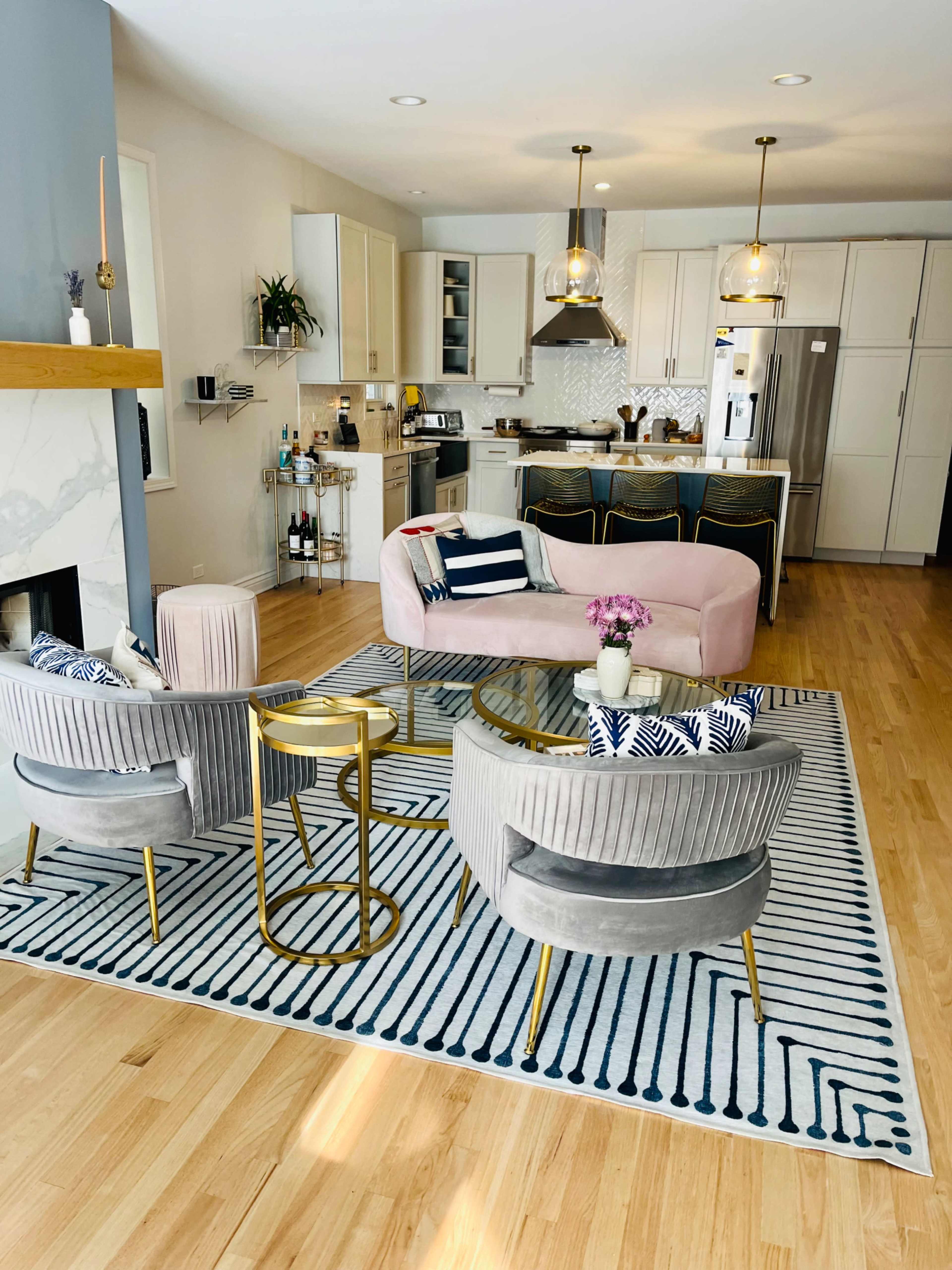 A modern living room with a pink sofa, two gray armchairs, a round gold side table, and a kitchen in the background.