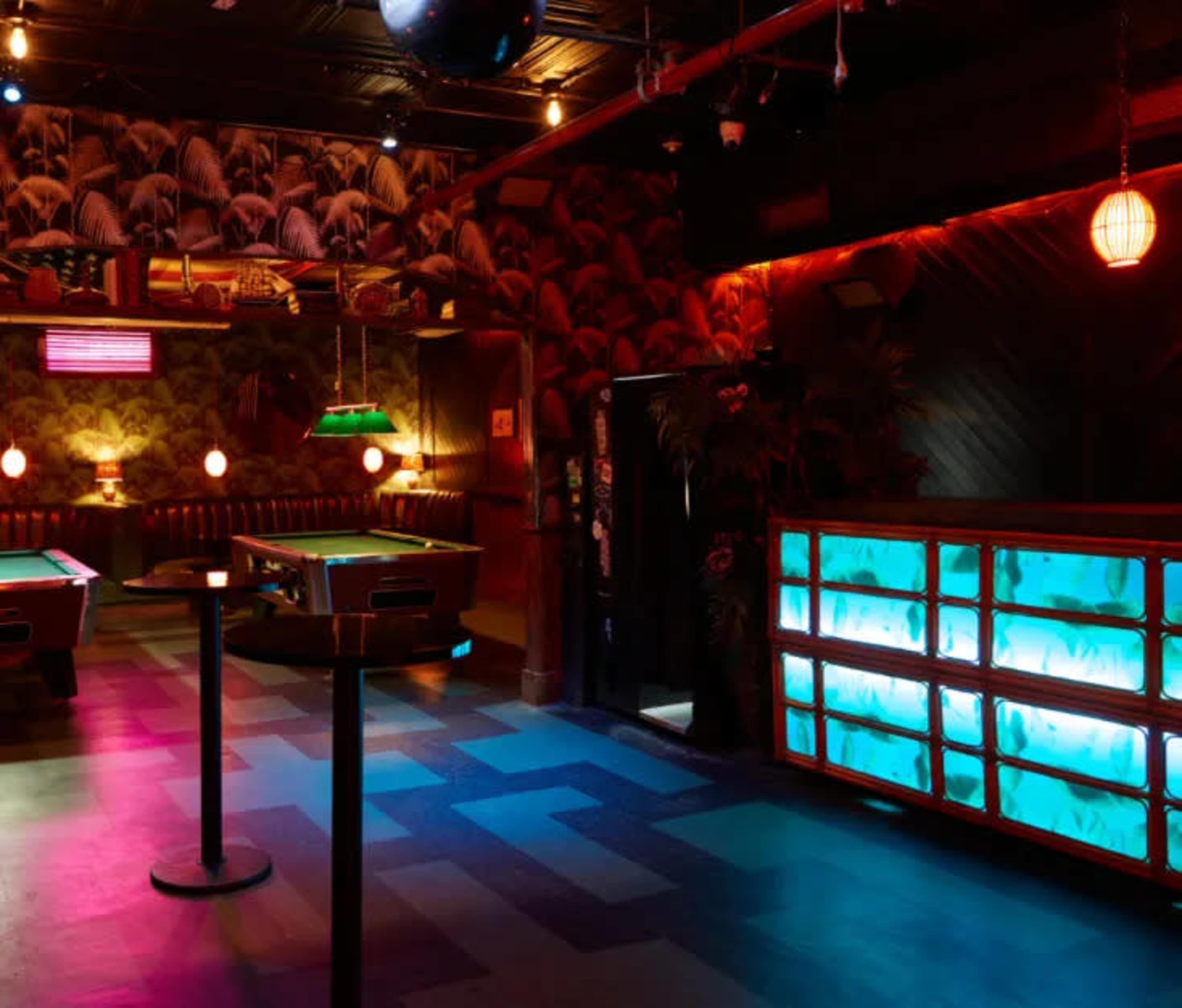 The image shows a dimly lit bar room featuring pool tables, decorative lighting, and tropical-themed wallpaper.