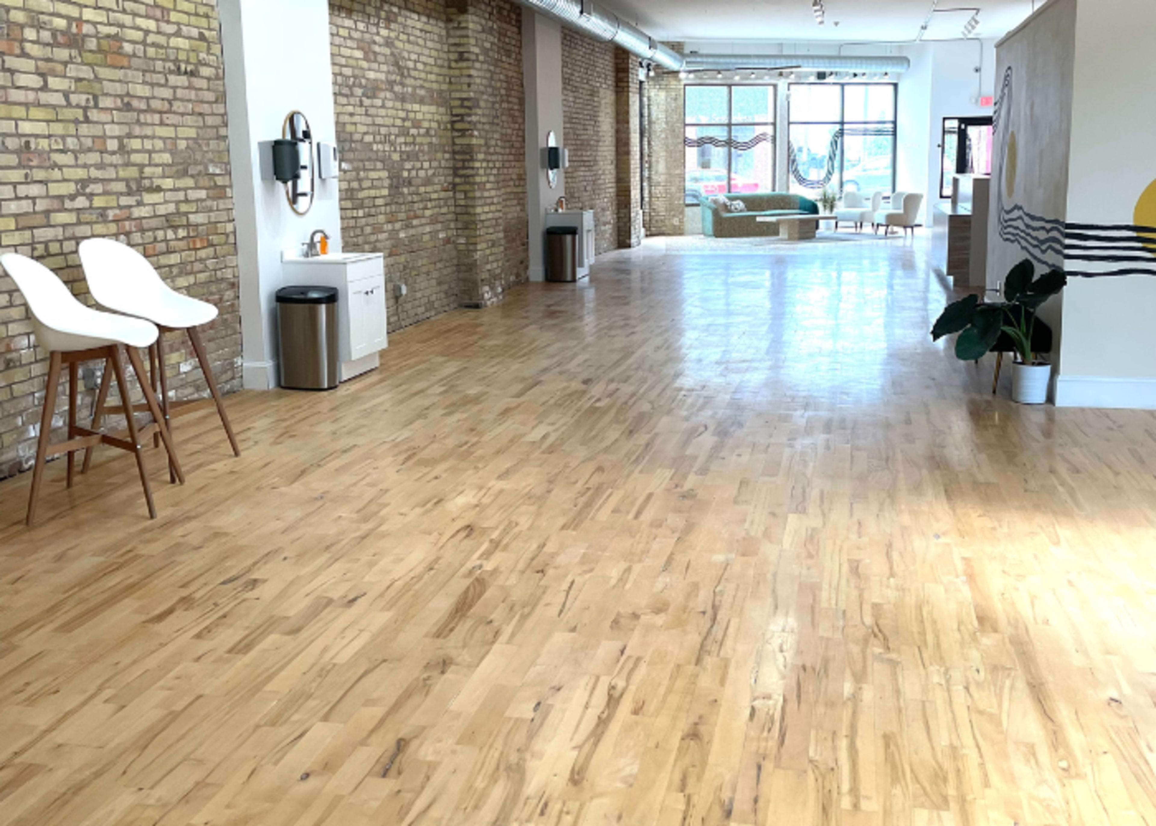 An empty, bright interior space with wooden flooring, exposed brick walls, and minimalist furniture.
