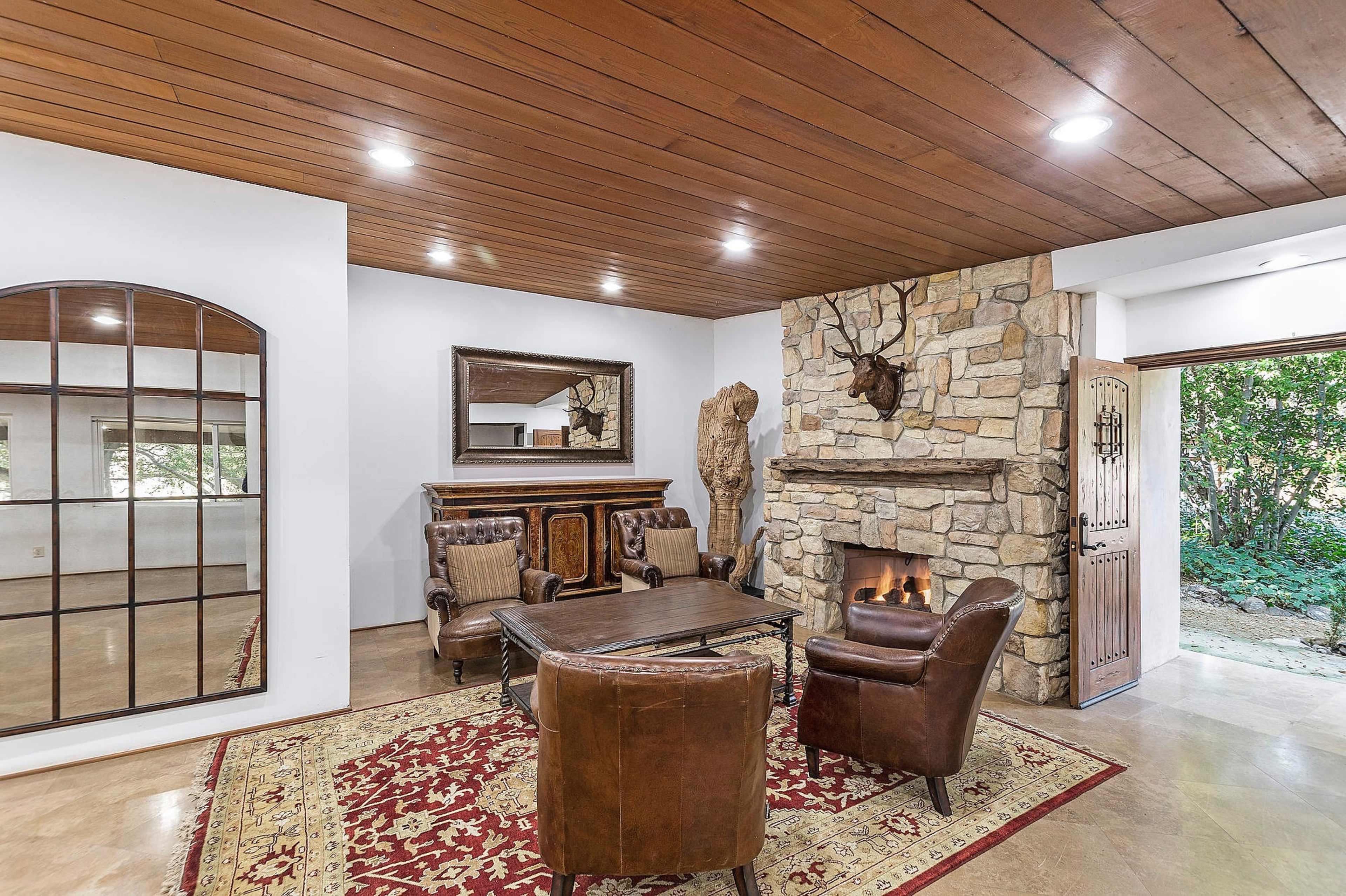 A cozy living room features a stone fireplace, a wooden ceiling, a large mirror, and leather armchairs arranged around a coffee table on a patterned rug.