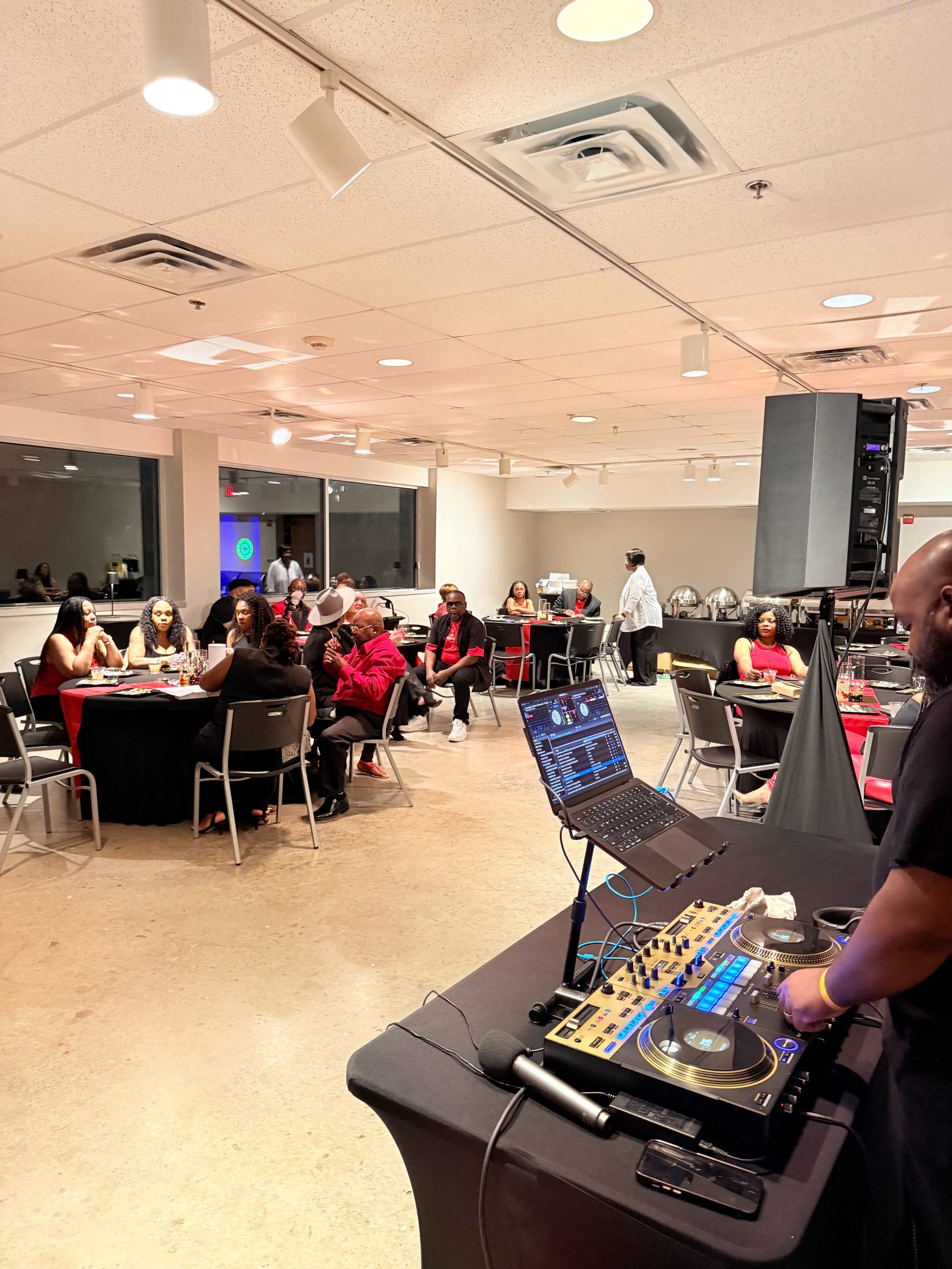 A DJ stands behind a mixer while guests sit at tables dressed with red and black linens, socializing in a well-lit event space.