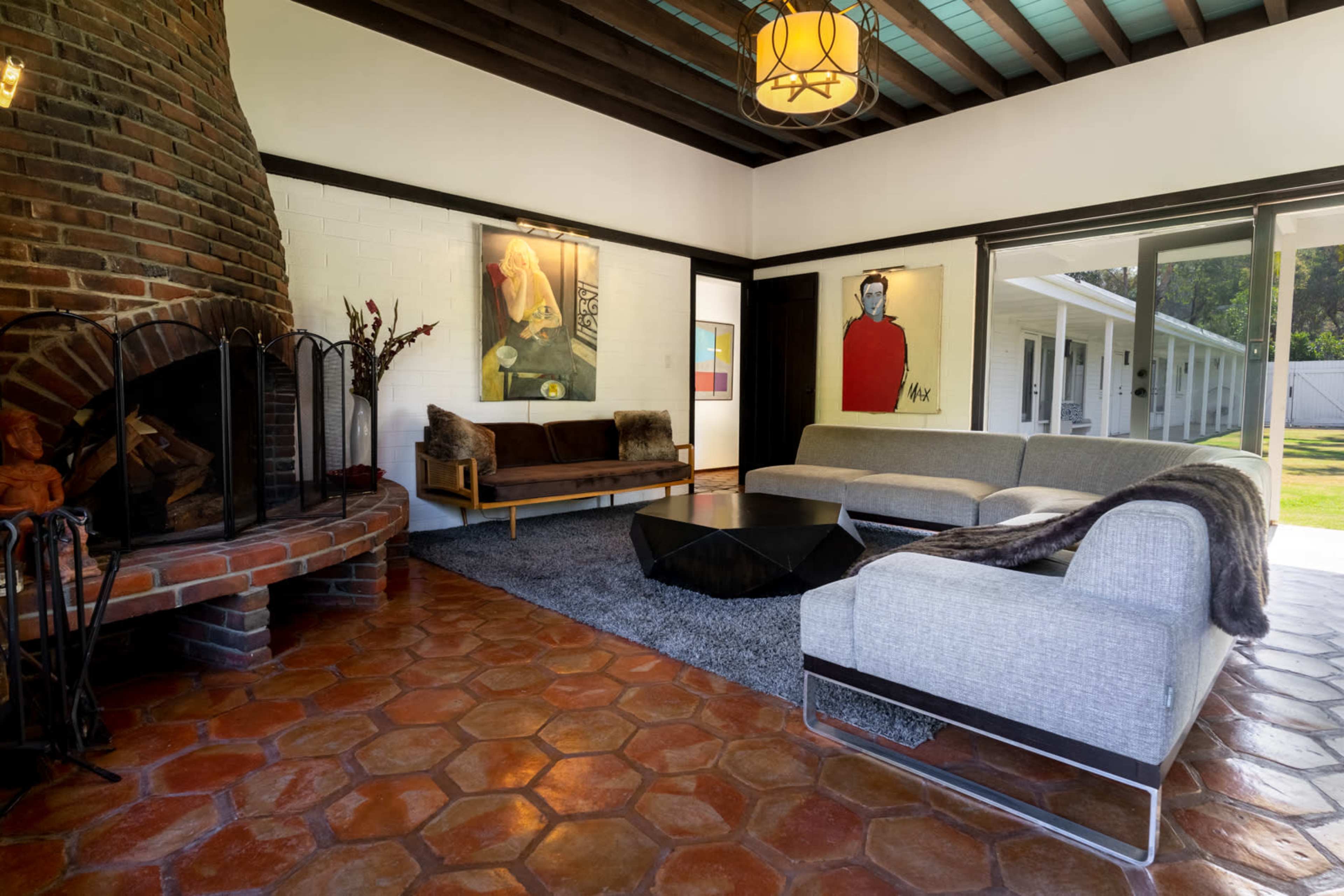 A spacious living room featuring a brick fireplace, two sofas, and colorful artwork on the walls, with large windows overlooking an outdoor area.