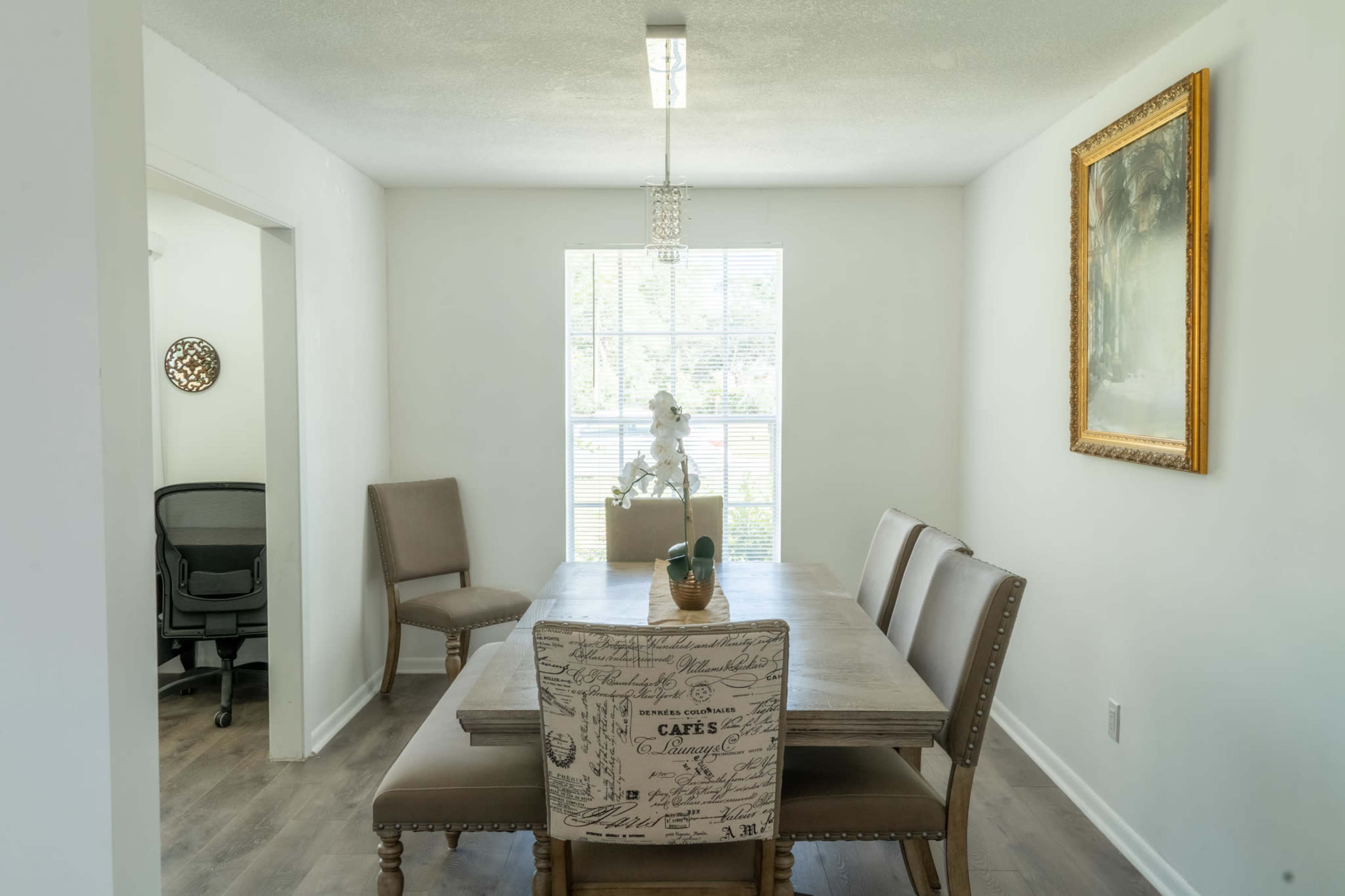 A light-filled dining area featuring a wooden table surrounded by five chairs, with a chair featuring a patterned cover, a piece of artwork on the wall, and an office space visible through an open doorway.