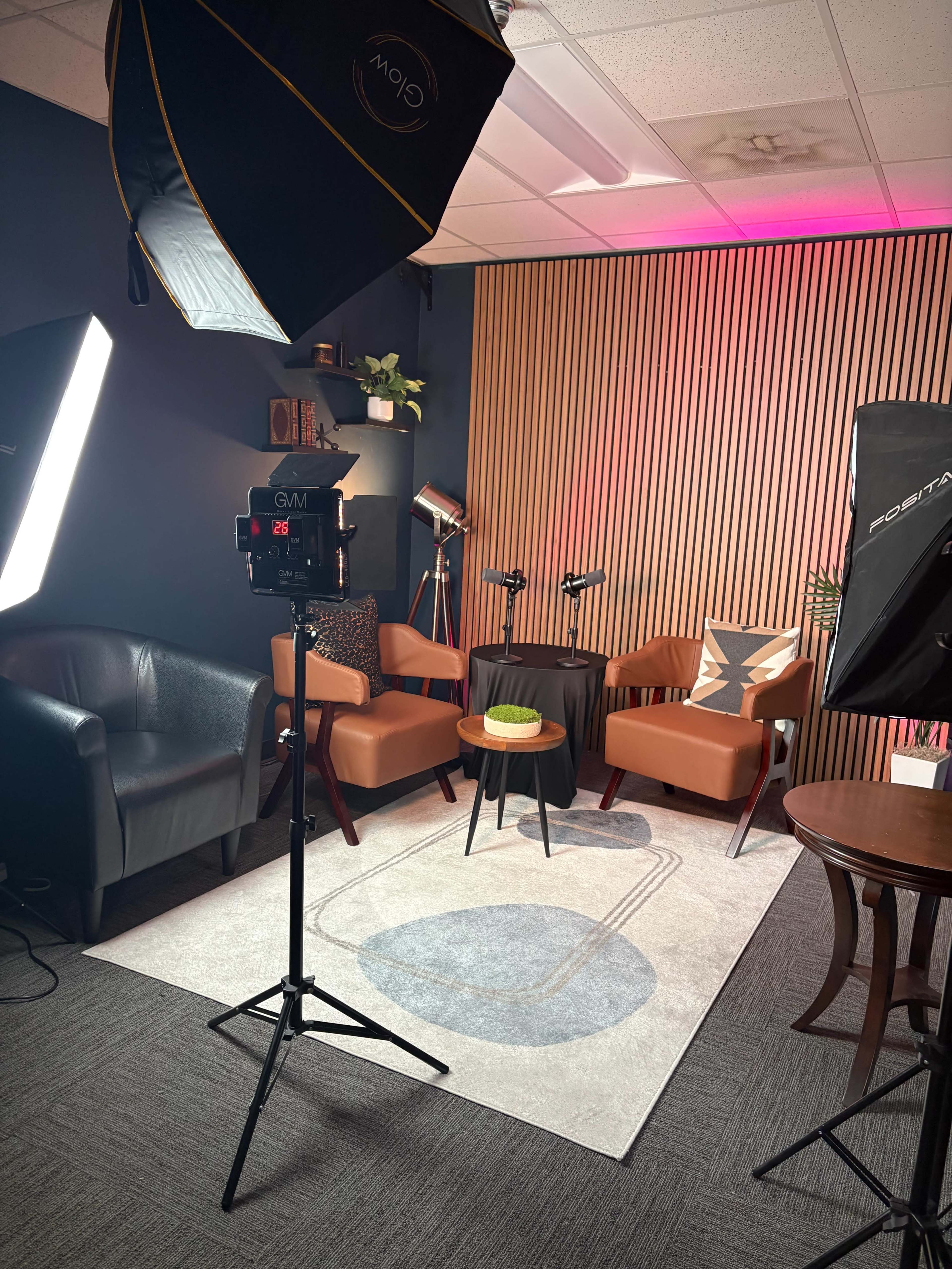The image shows a neatly arranged indoor studio setup featuring two brown chairs, a small round table, and lighting equipment against a backdrop of wooden paneling.