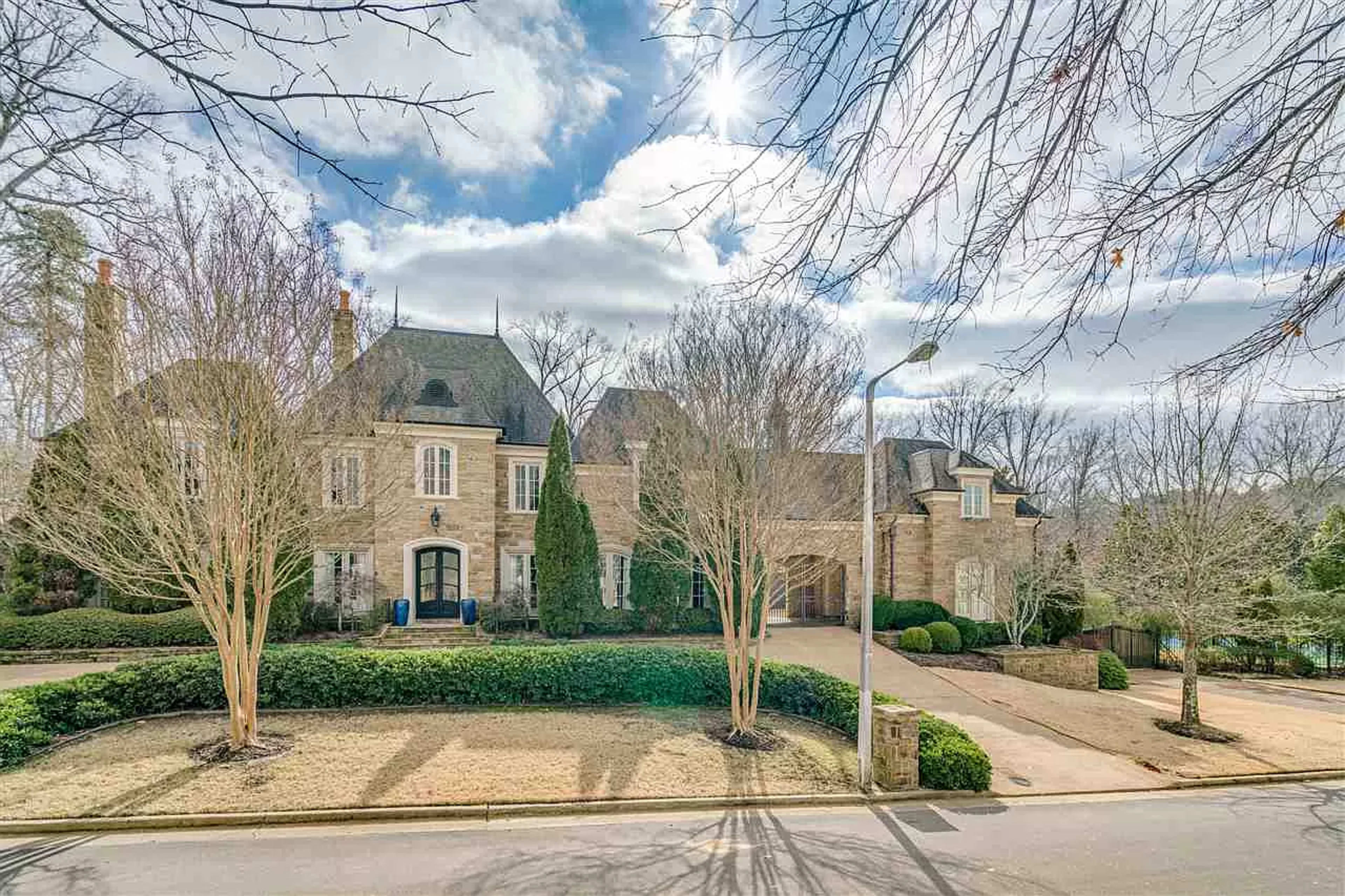 A large stone mansion is set back from the street, surrounded by neatly trimmed bushes and bare trees under a partly cloudy sky.