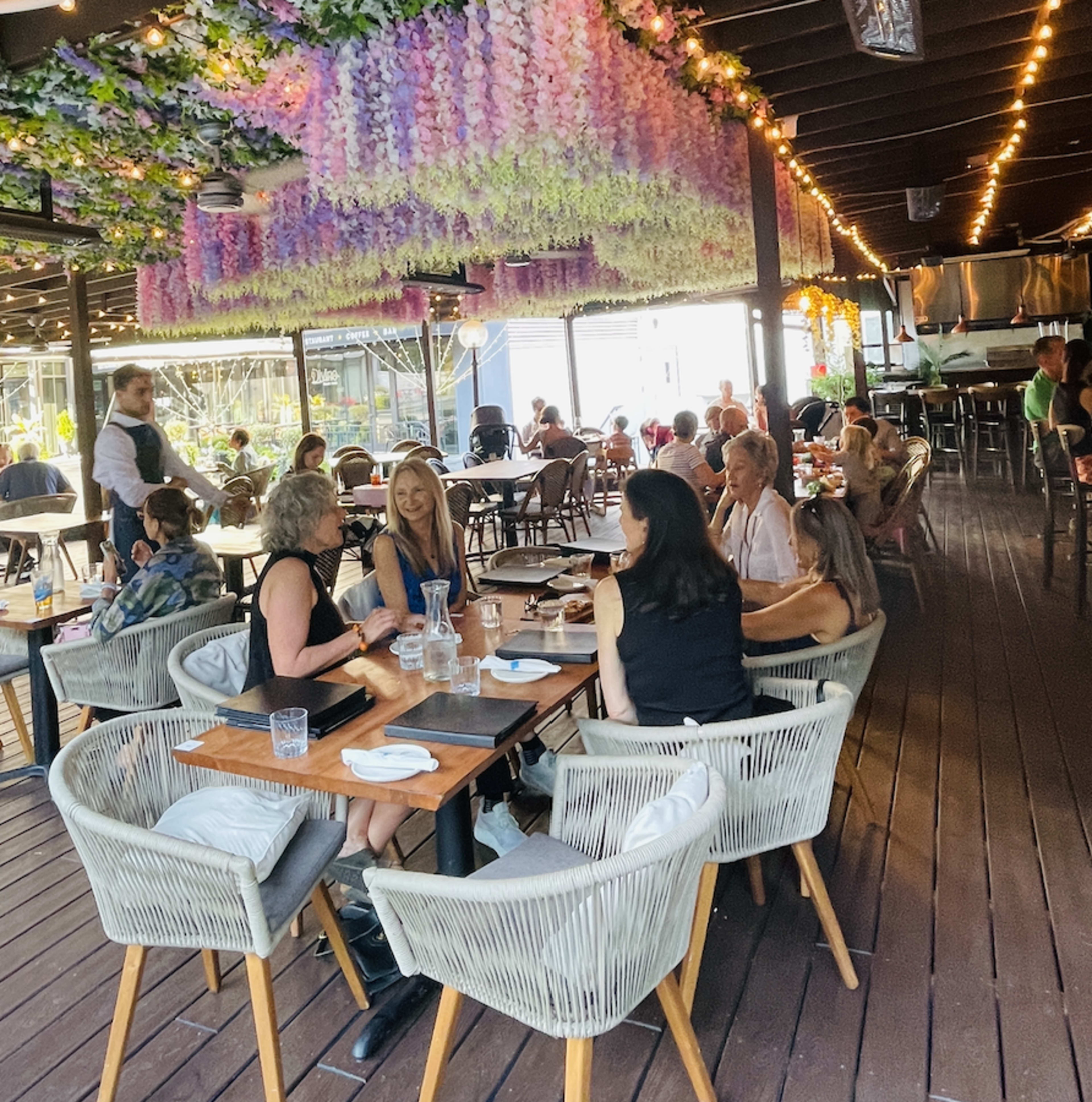 The restaurant features a lively outdoor seating area with several diners enjoying their meals under floral decorations and string lights.