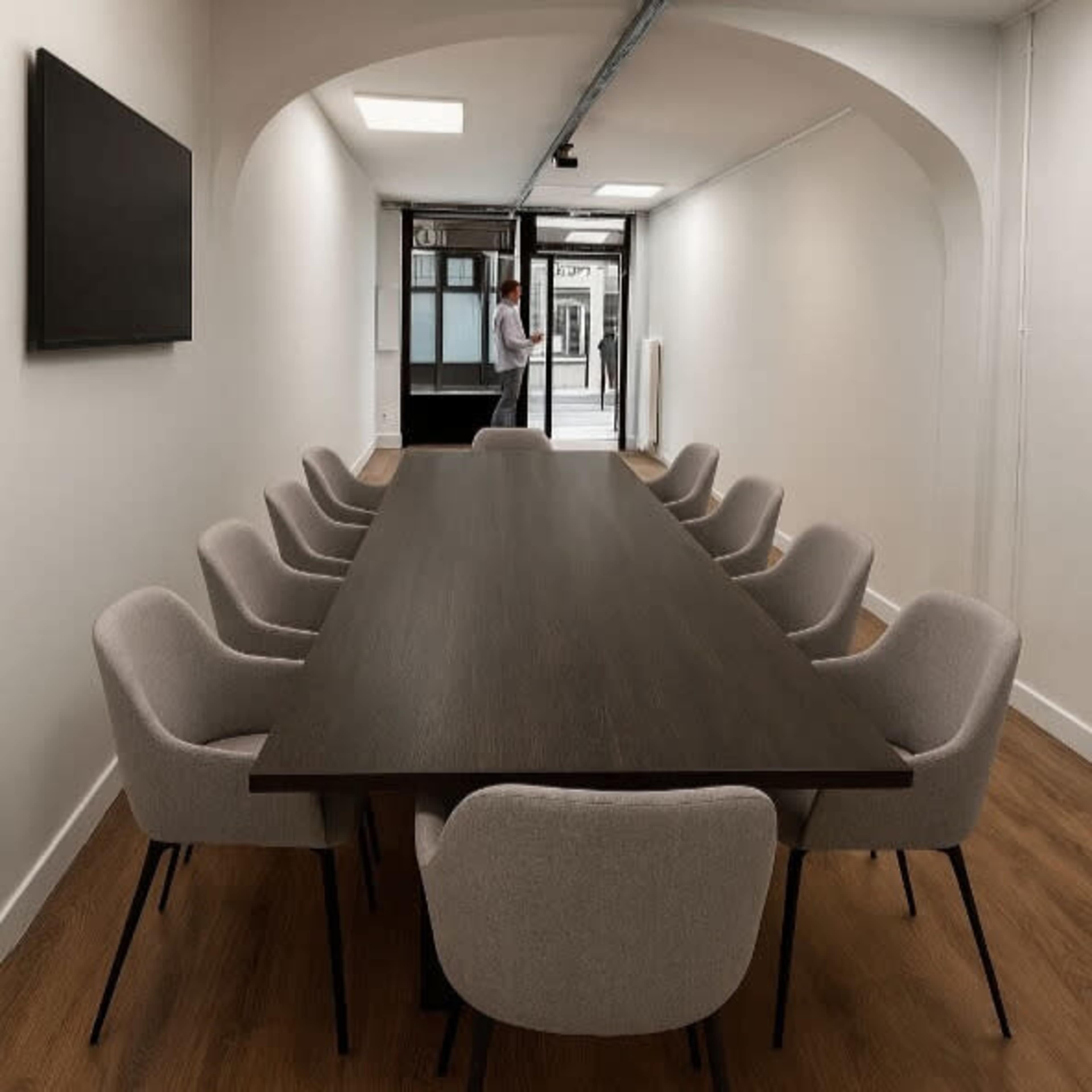A long, dark wooden conference table is surrounded by light gray upholstered chairs in a well-lit, modern meeting room.