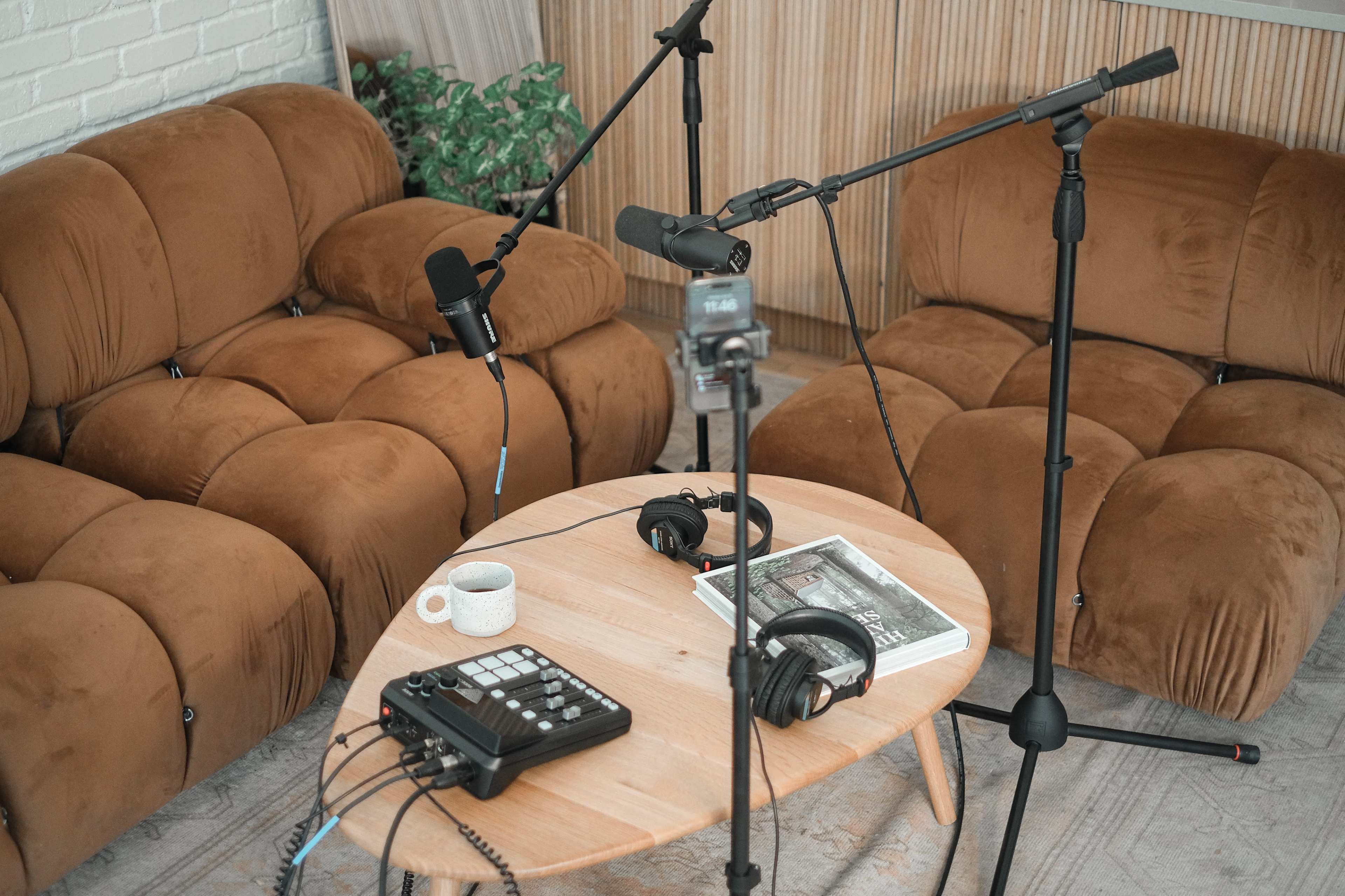 A small table with a mixer and headphones is surrounded by two brown sofas and microphone stands in a recording setup.