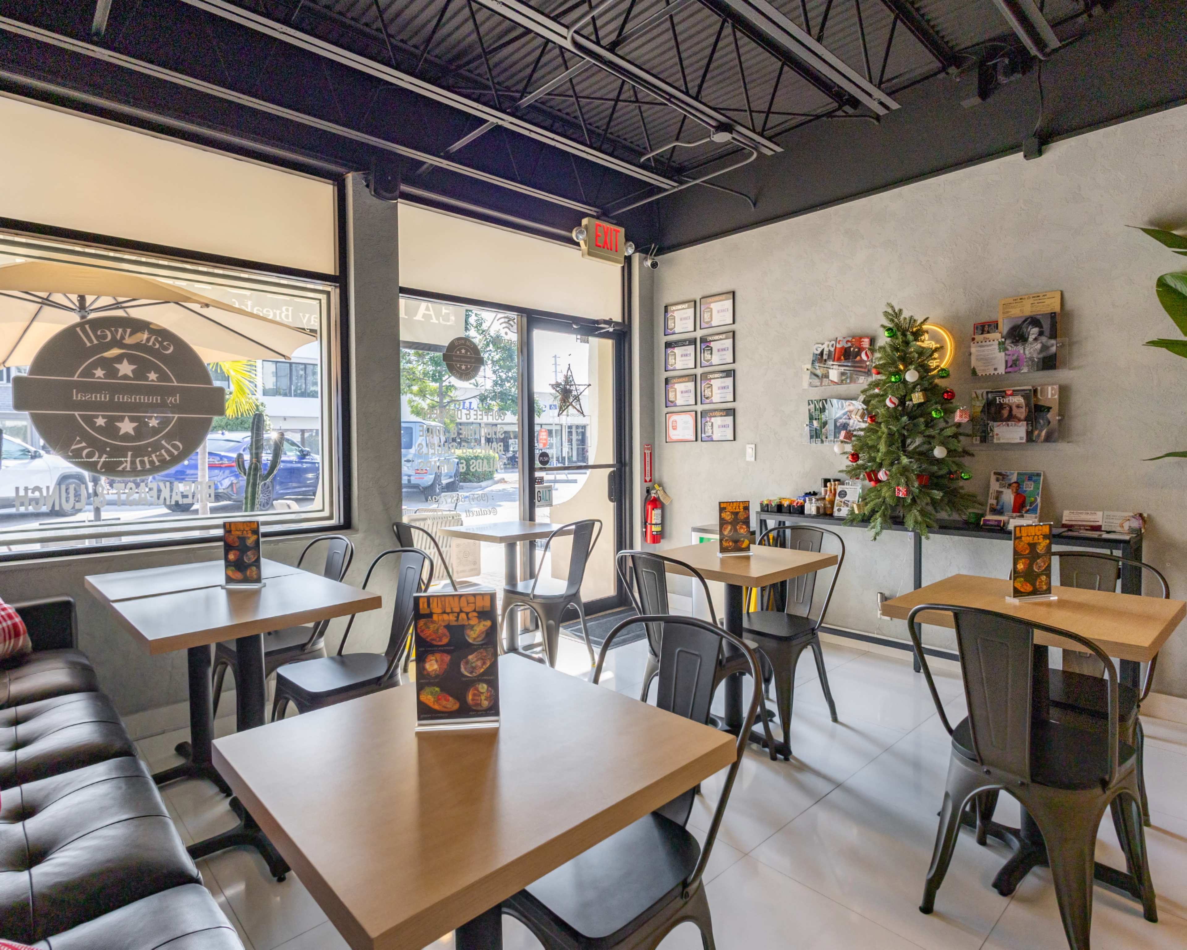 The image shows a modern cafe interior with several tables, metal chairs, a decoratively adorned Christmas tree, and large windows allowing natural light to enter.