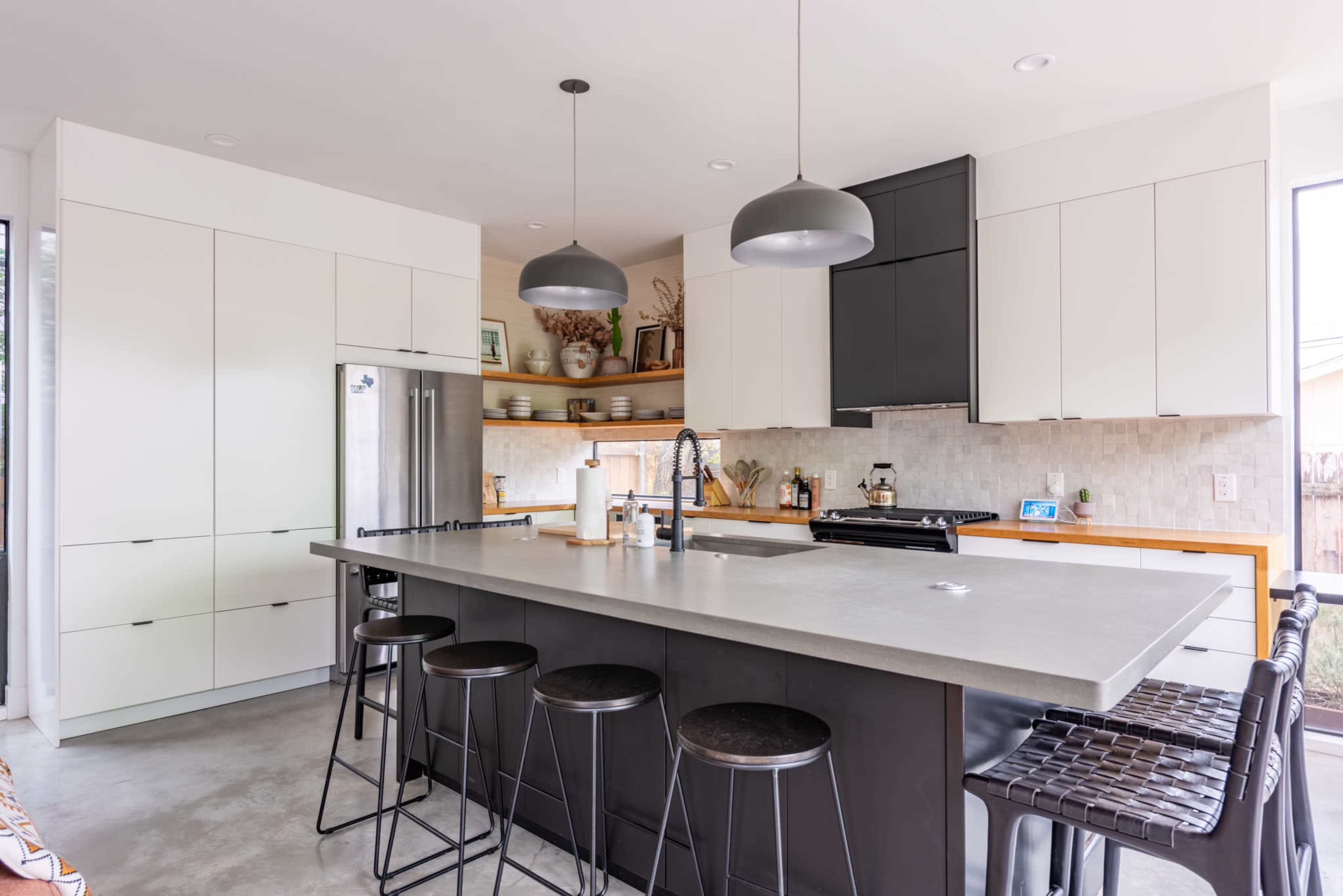 The kitchen features a large concrete island with black stools, stainless steel appliances, and a mix of white and black cabinetry.