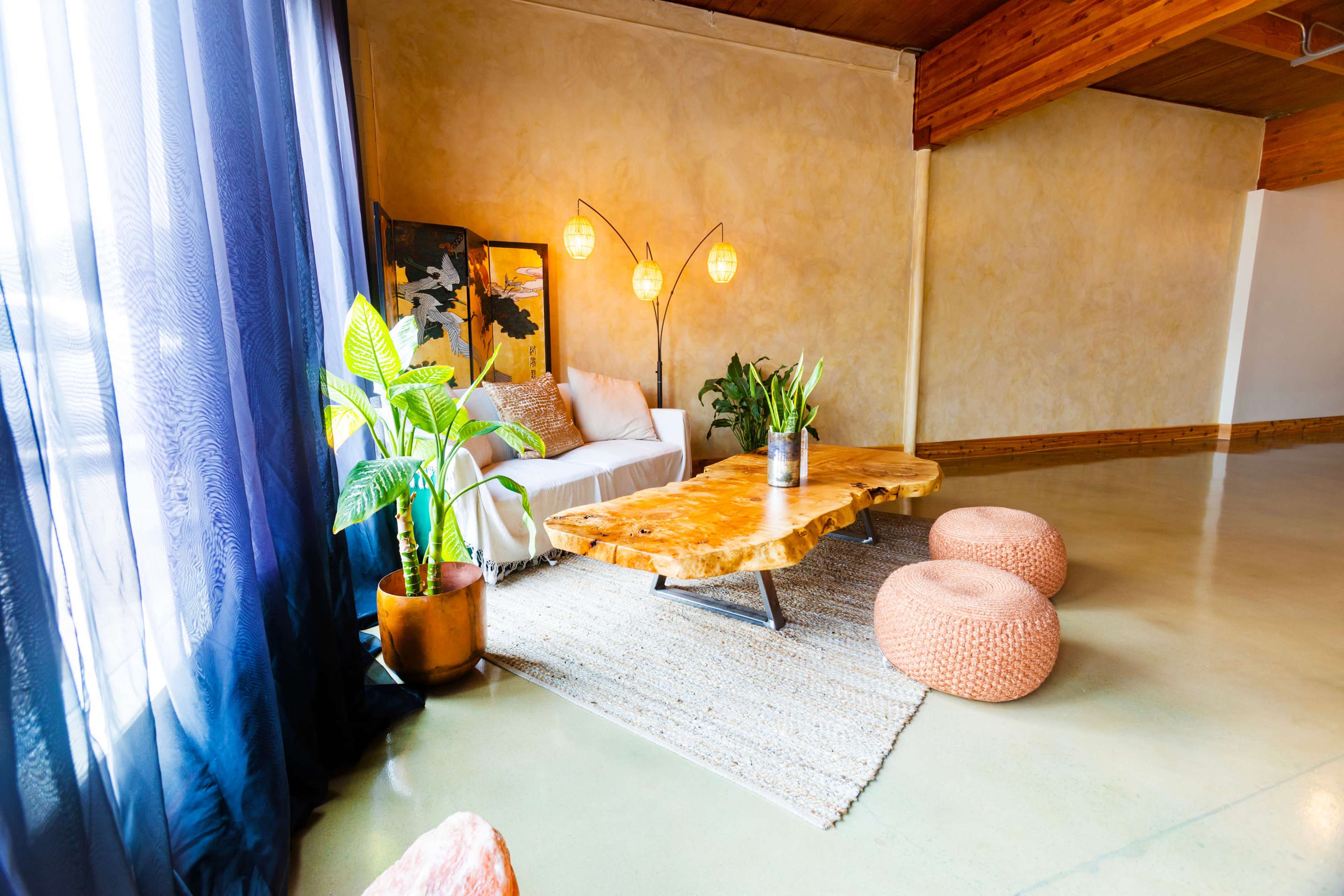 A modern interior living space featuring a wooden coffee table, a couch with white cushions, decorative plants, and round poufs on a textured rug.