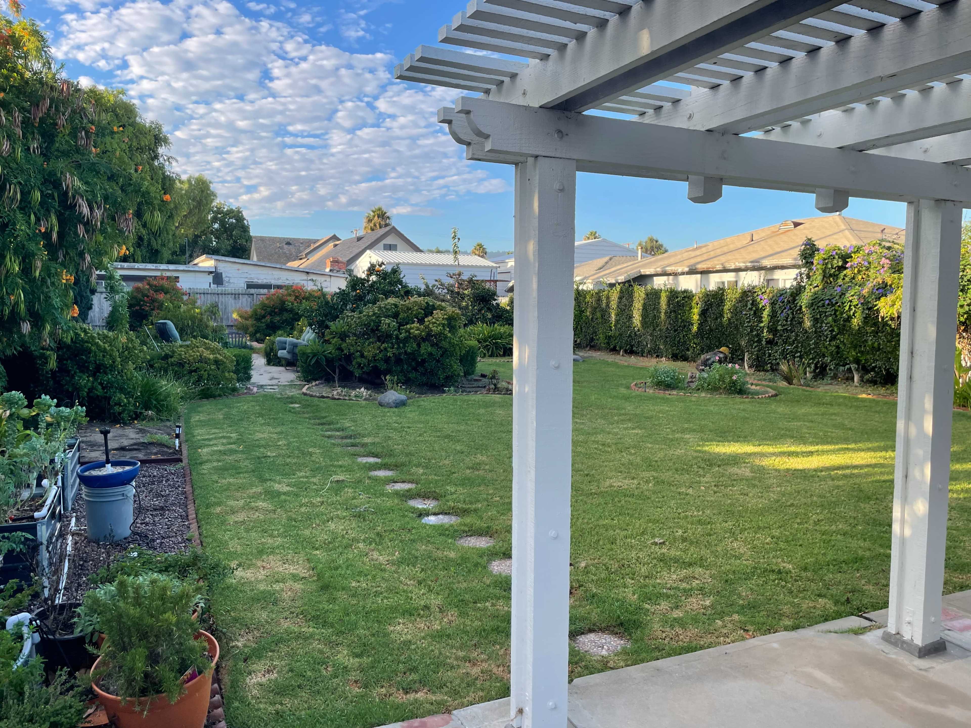 The image shows a backyard with a well-maintained lawn, garden beds, and a pergola in the foreground.