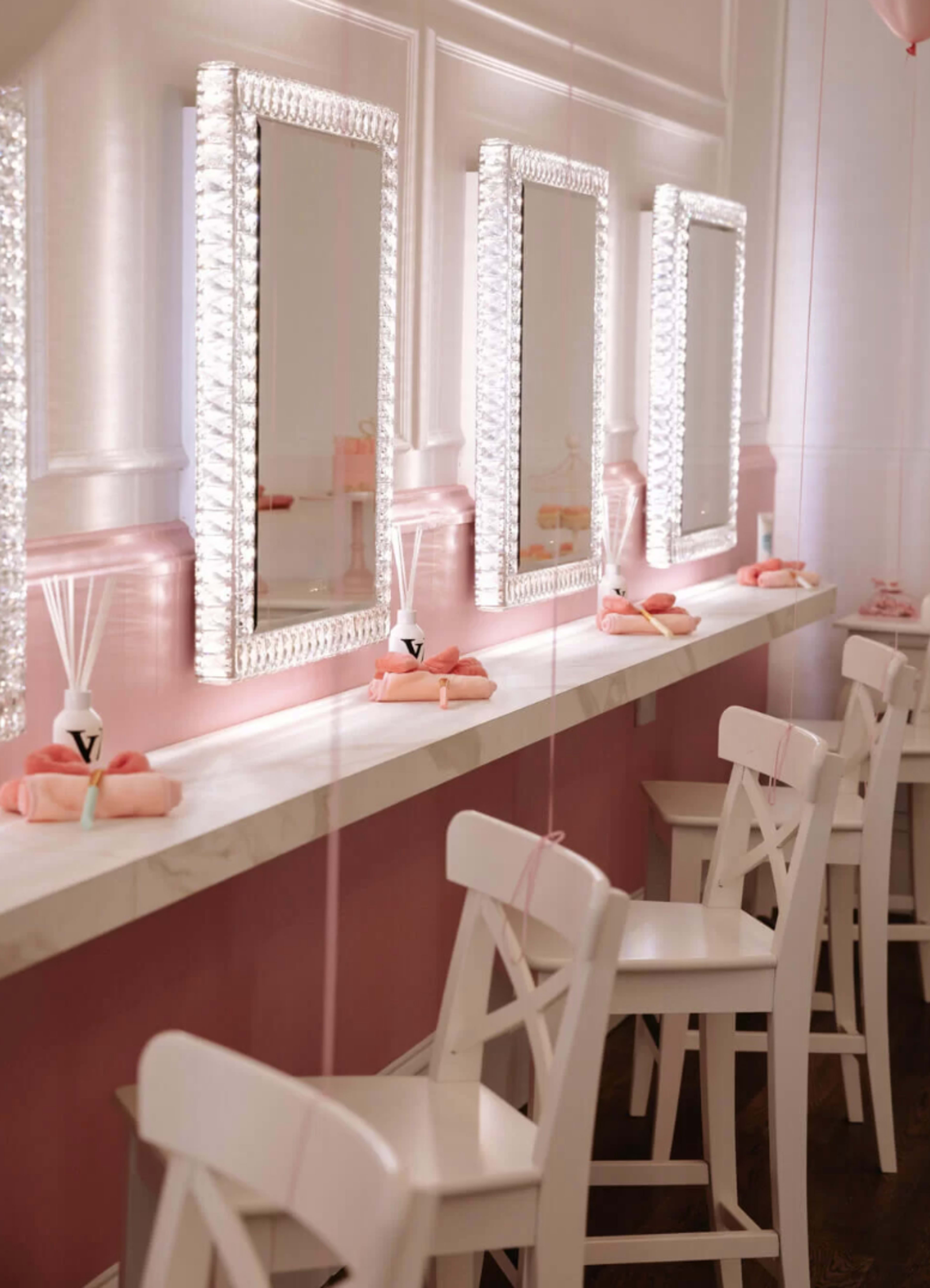 A row of four illuminated mirrors is positioned above a marble countertop with pink decor and white chairs.