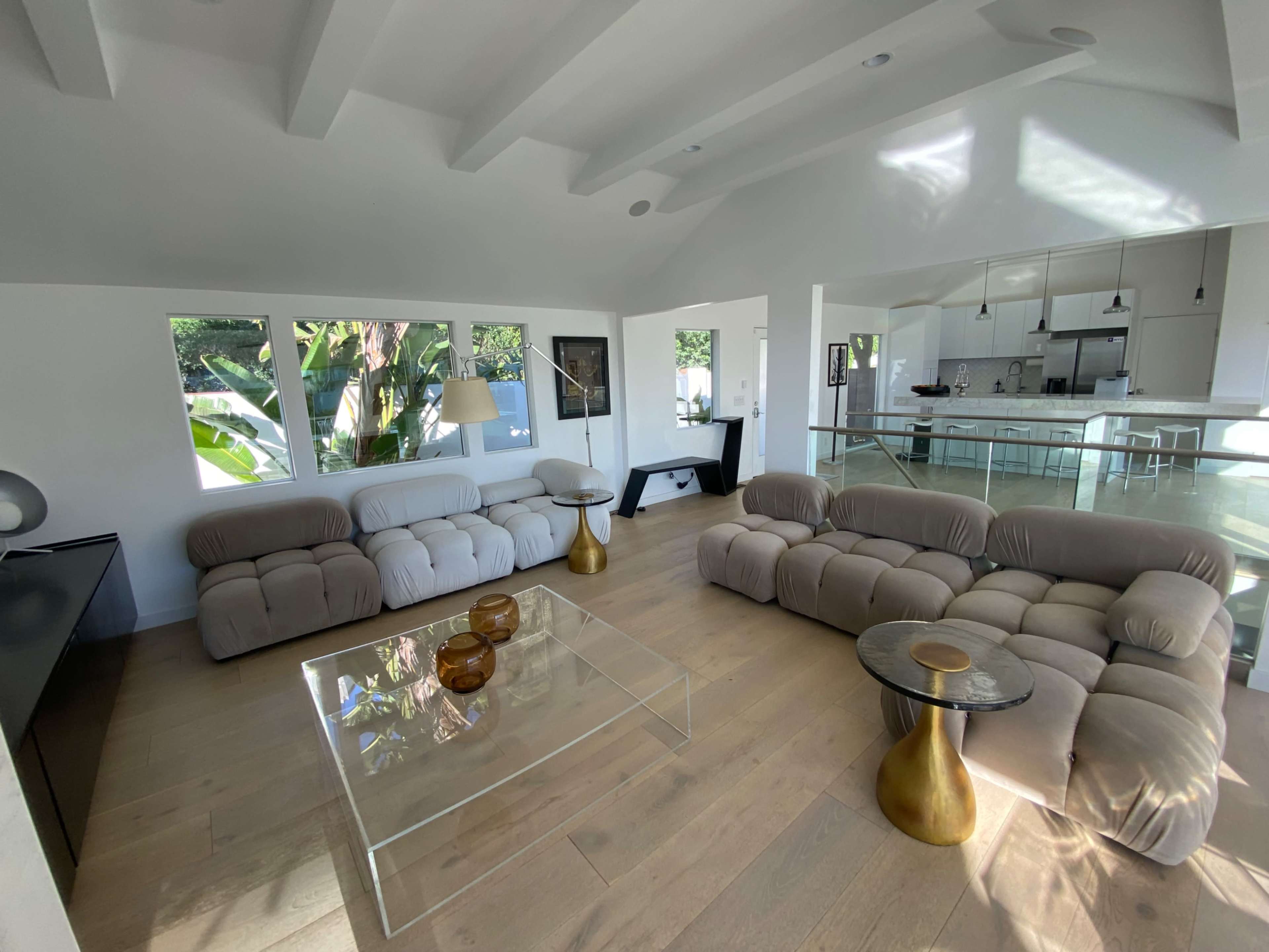 The image shows a modern living room featuring two beige sofas, a glass coffee table with gold accents, and large windows allowing natural light in.