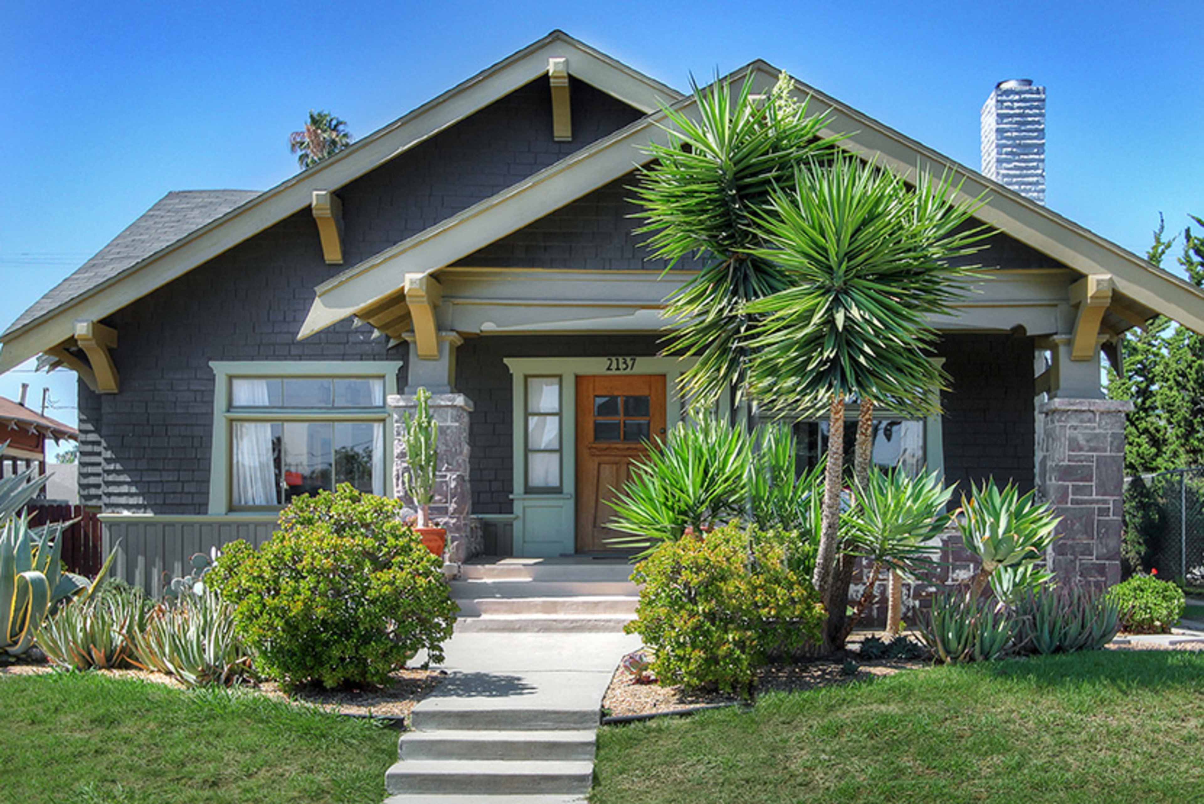 A gray bungalow with a front porch and two palm-like plants flanking the entrance.