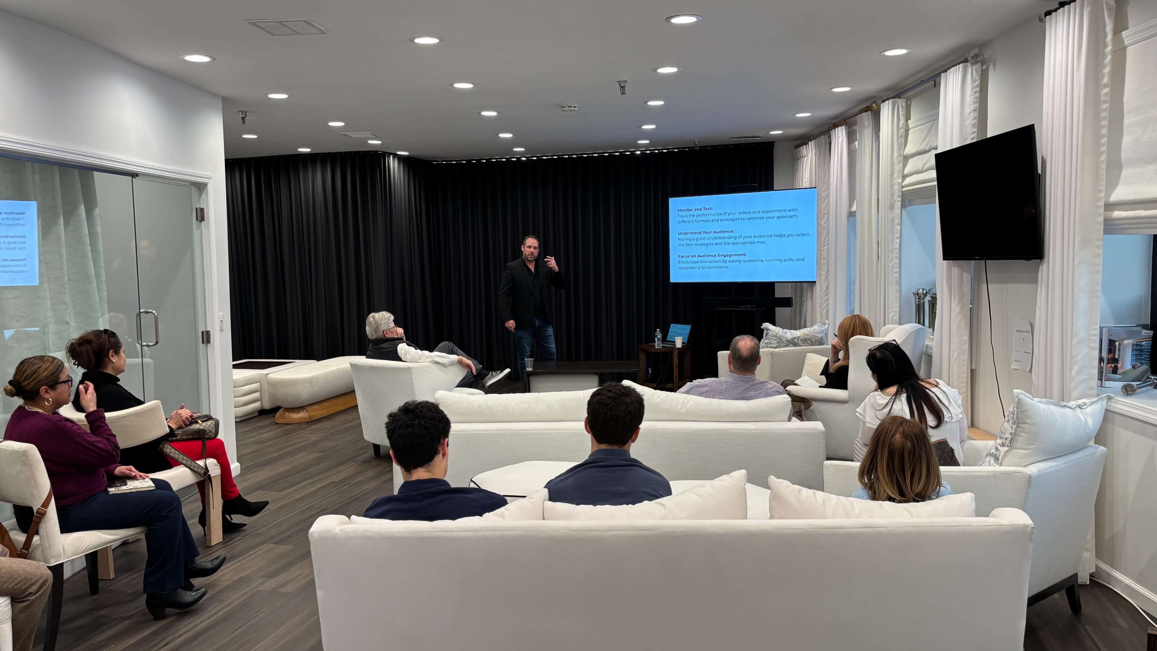 A speaker presents information to an audience seated on sofas in a modern room with large windows and a screen displaying slides.