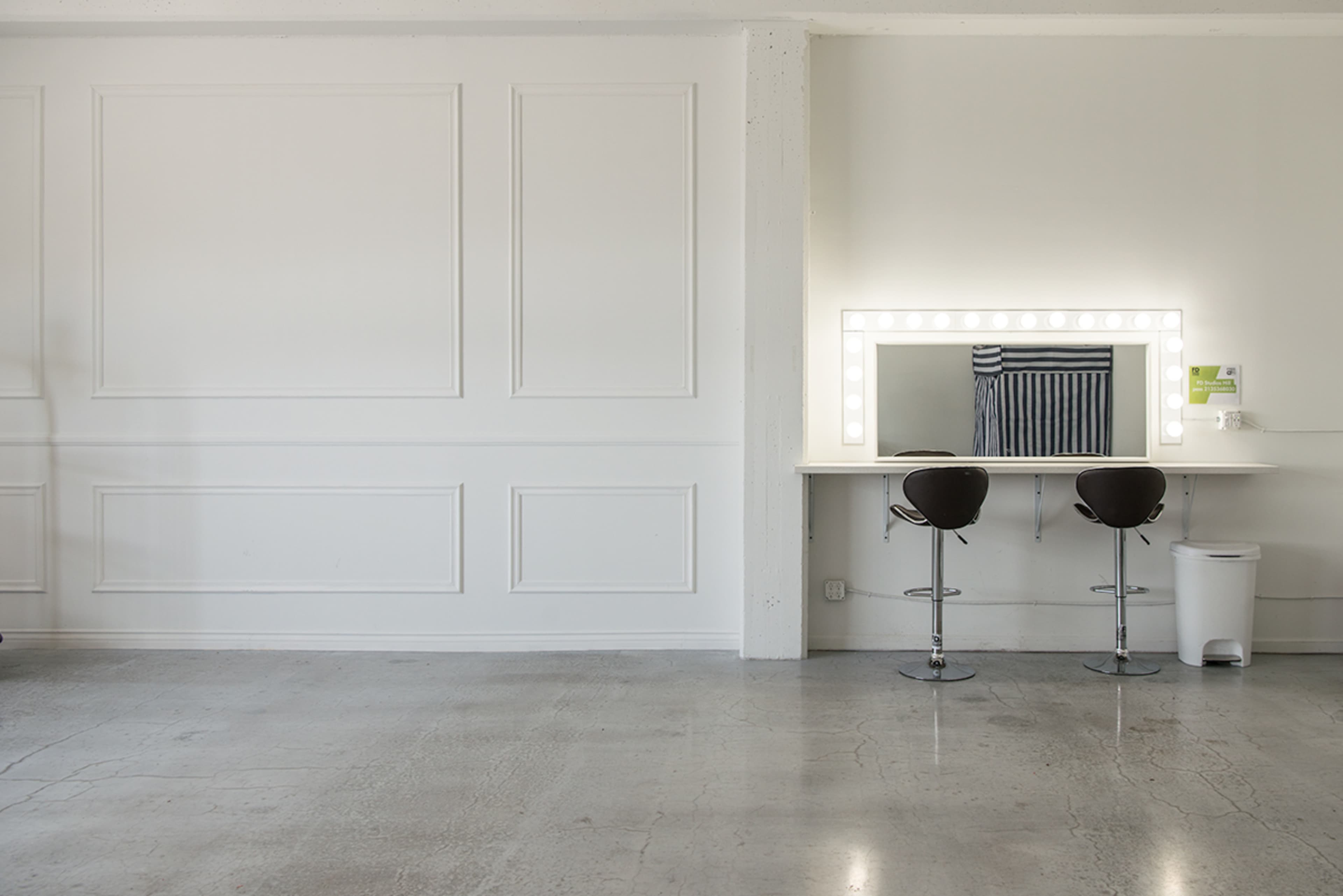 The image shows a minimalist room with a white wall, two bar stools facing a vanity mirror surrounded by lights, and a small trash bin nearby.