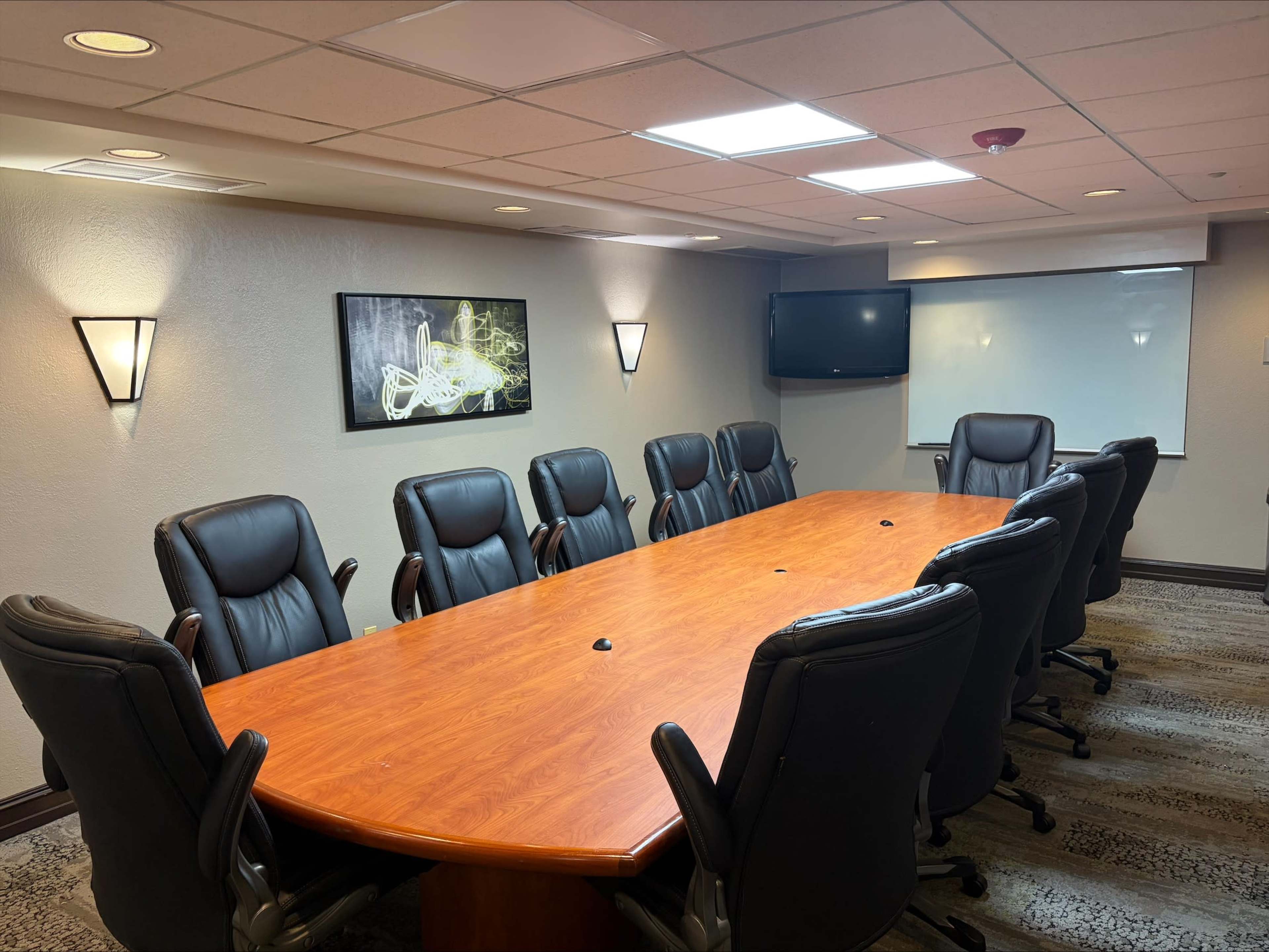 A conference room features a long wooden table surrounded by thirteen black leather chairs, with a monitor and whiteboard on the wall.