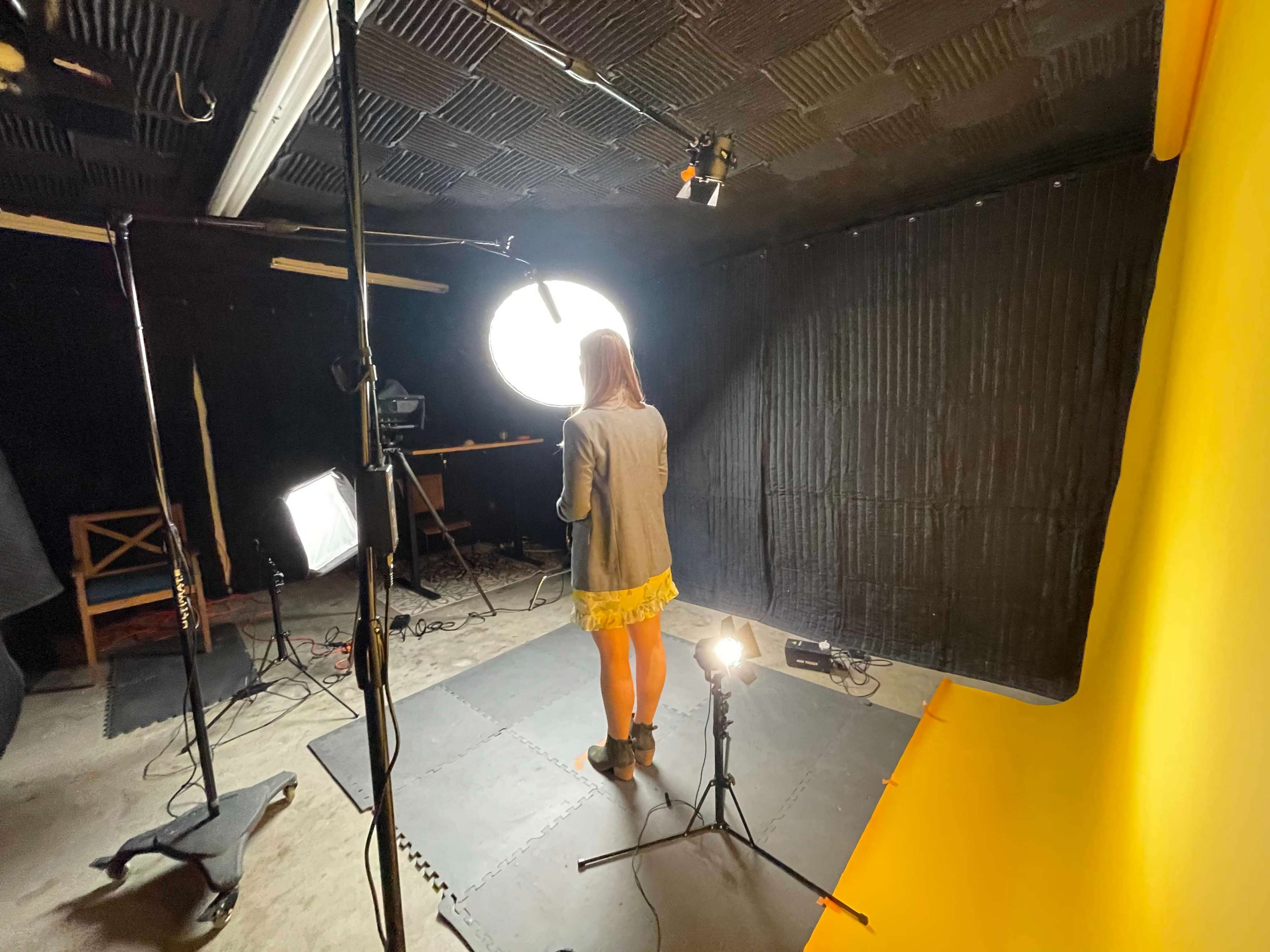A person stands in a photography studio with yellow backdrop, surrounded by studio lights and equipment.