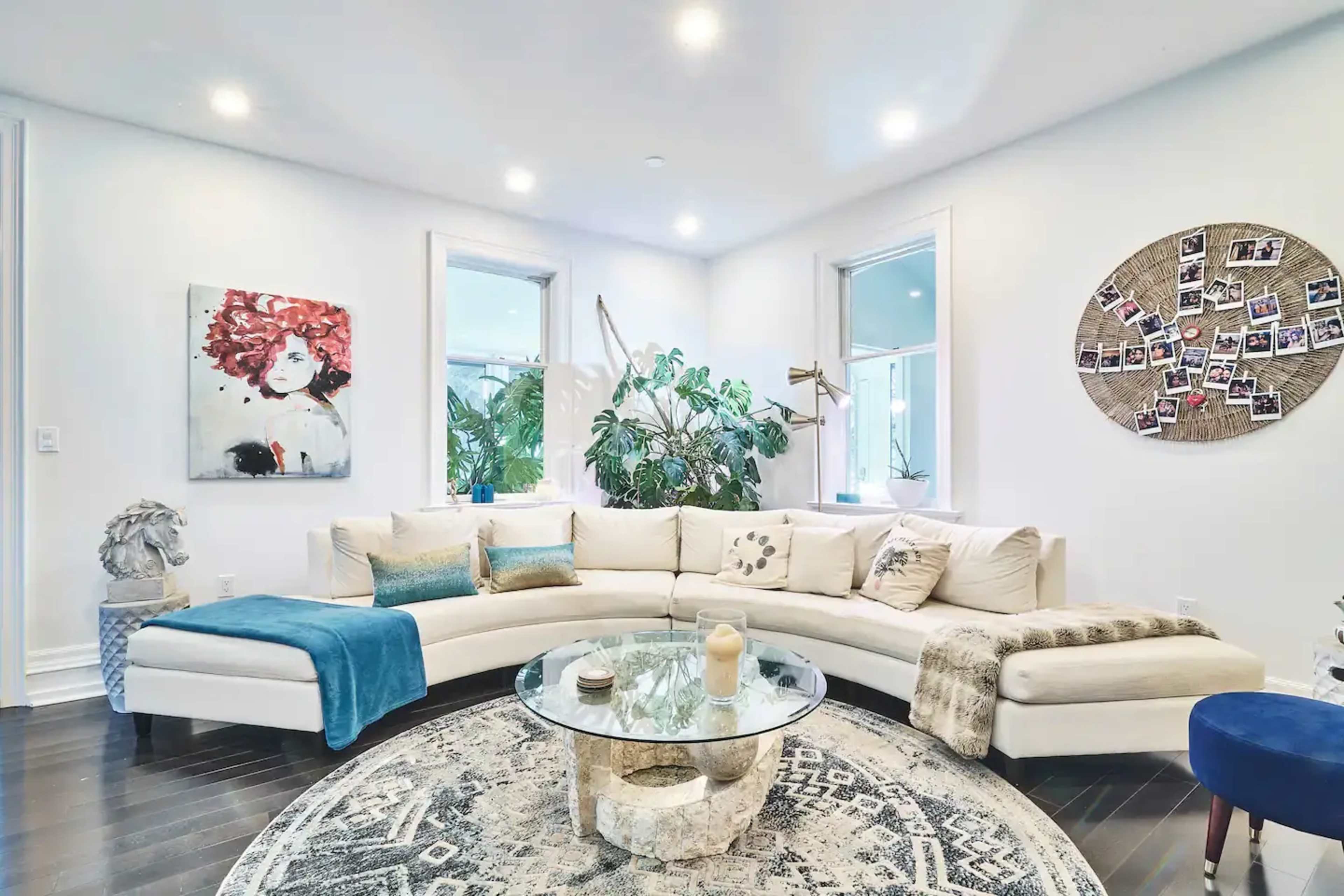 A spacious living room features a curved white sectional sofa, a round glass coffee table, and large windows surrounded by plants.