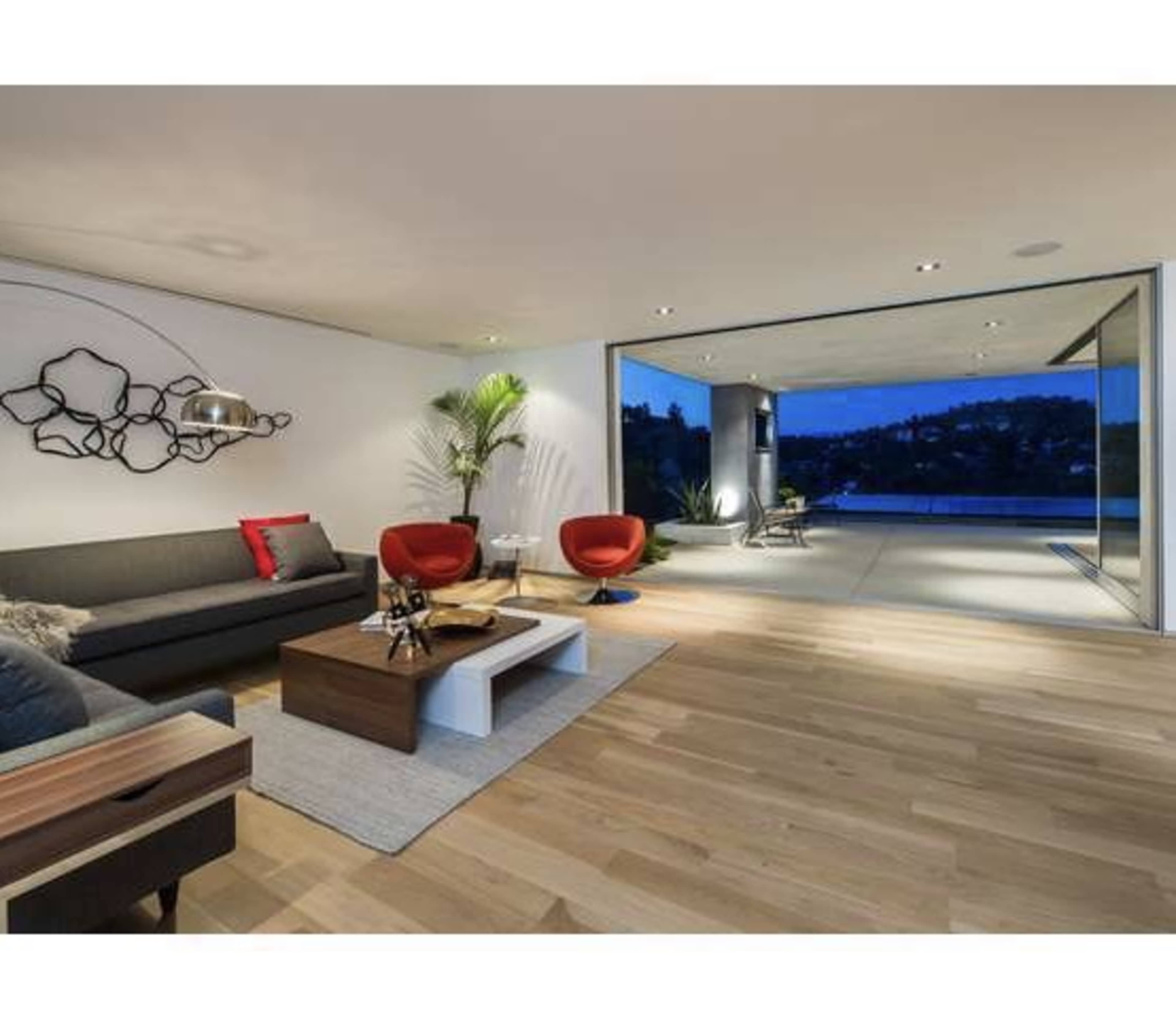 The image shows a modern living room with a dark sofa, two red chairs, and large glass doors opening to an outdoor view.