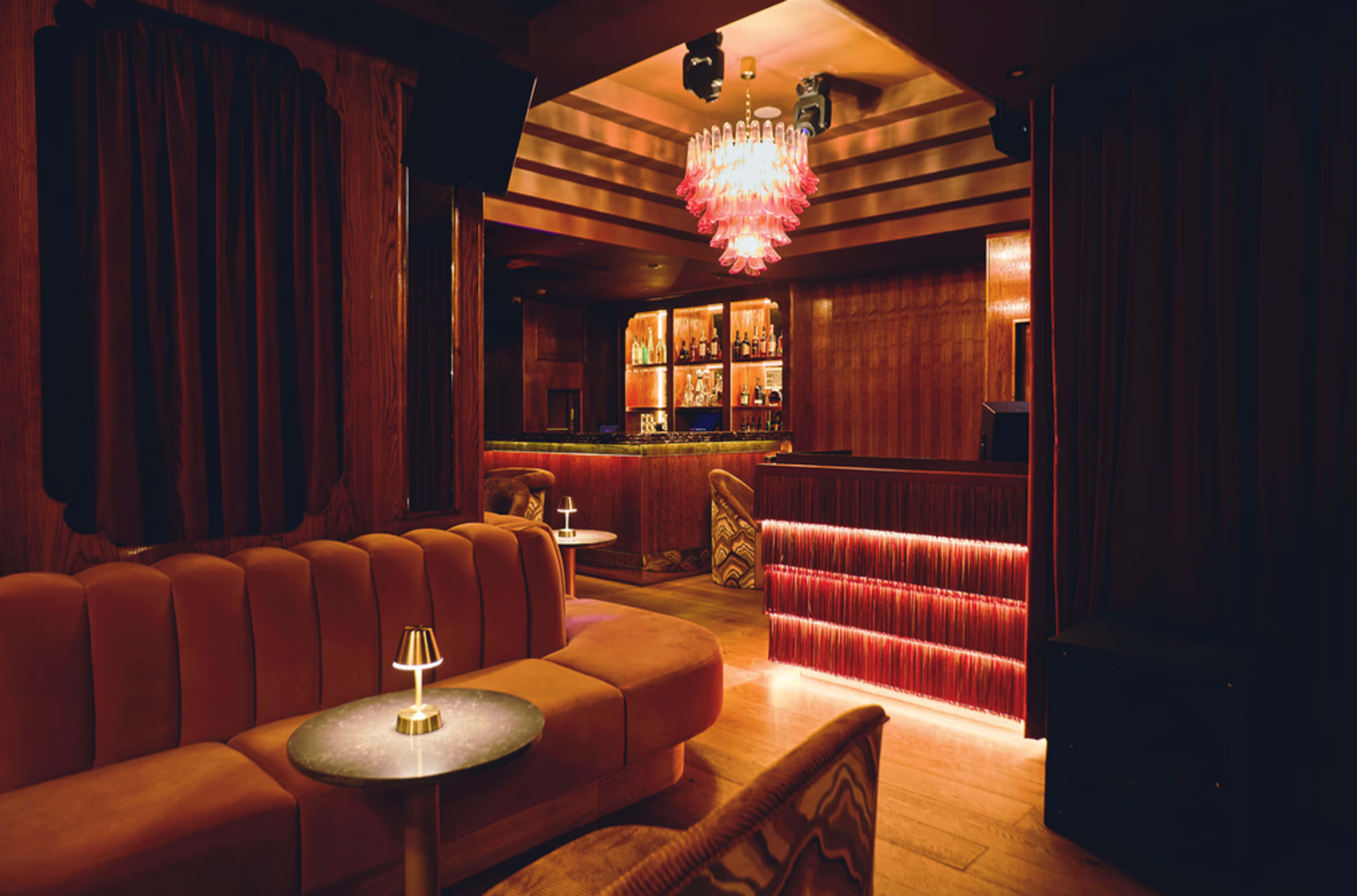 The image shows a cozy lounge with a curved, plush seating area, a small round table, and a bar illuminated by warm lighting.