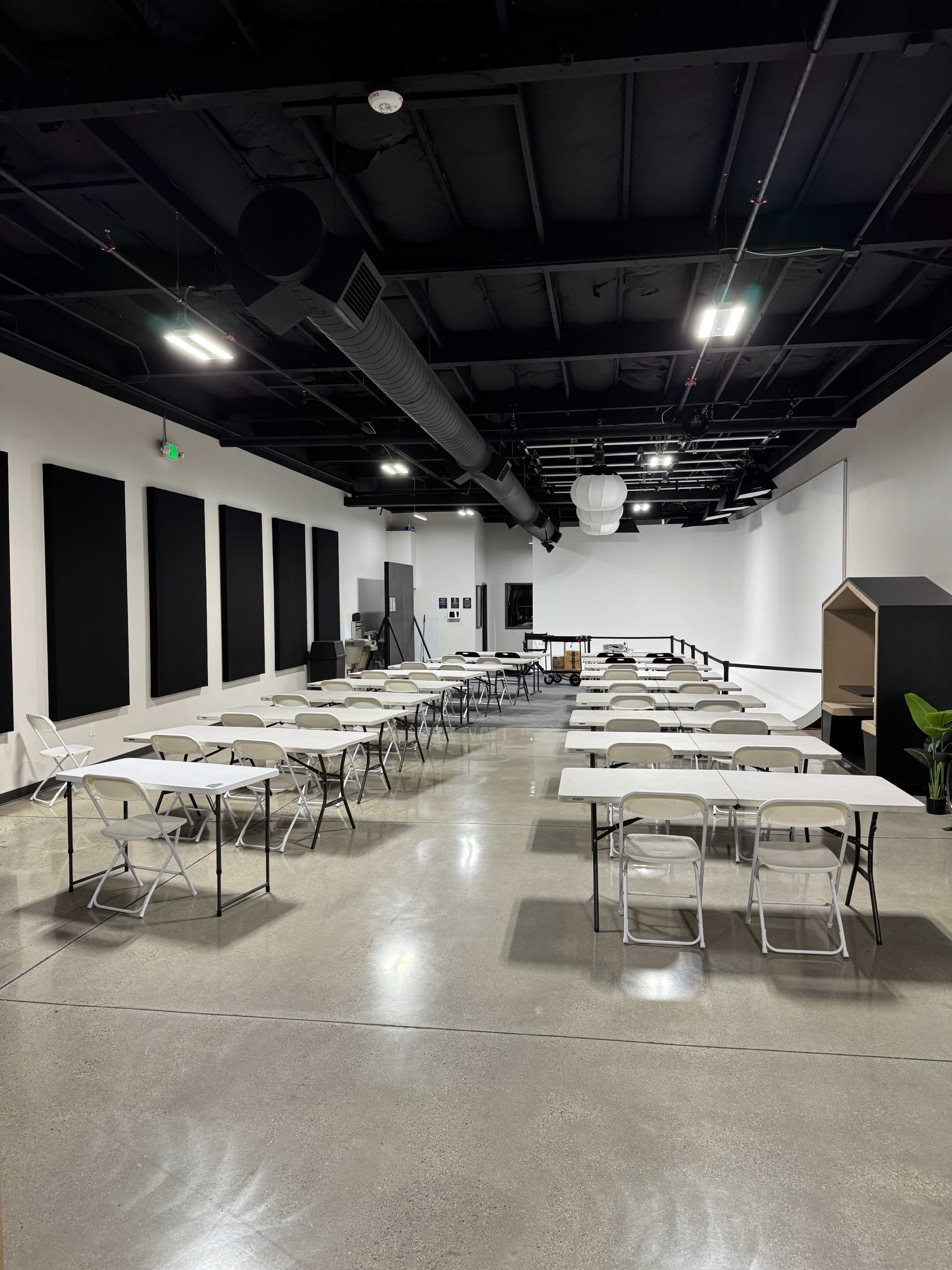 The image shows a spacious, well-lit room with white walls, featuring several tables and chairs arranged in a grid pattern.