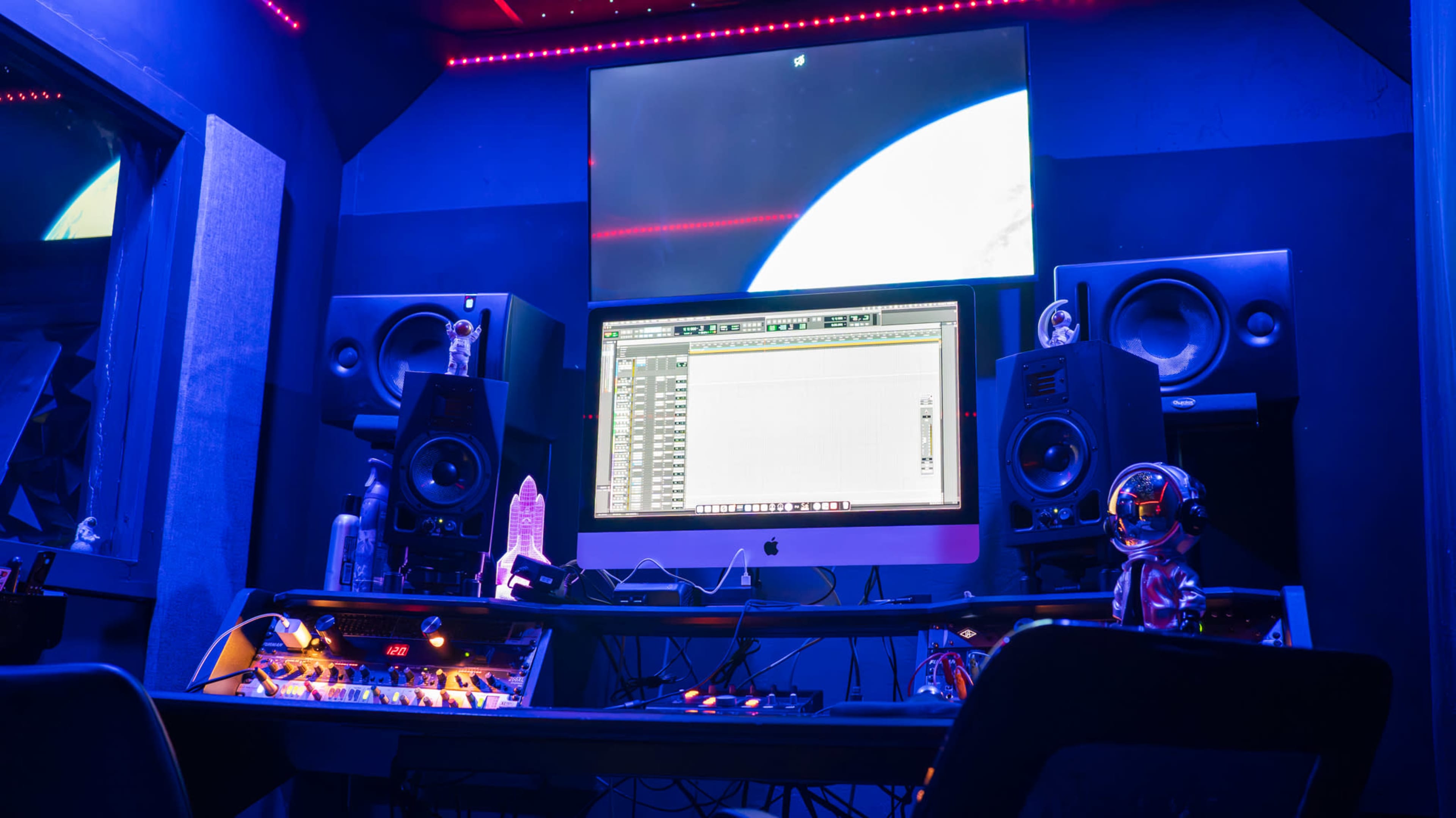 The image shows a dimly lit music studio featuring a large monitor, audio speakers, and various music production equipment arranged on a black desk.