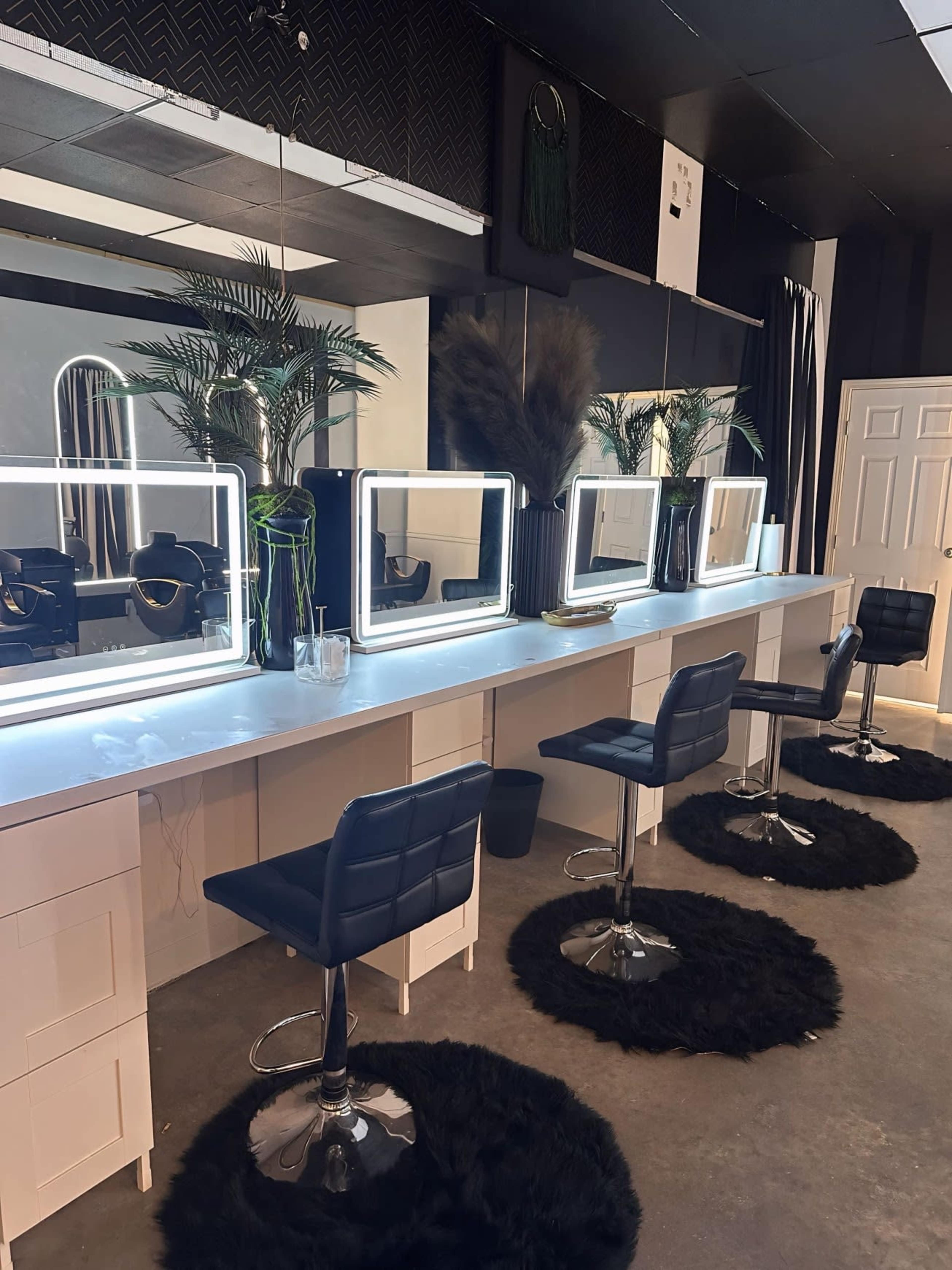 The image shows a modern beauty salon featuring several illuminated mirrors above sleek vanities, with black cushioned stools and circular black rugs on a concrete floor.