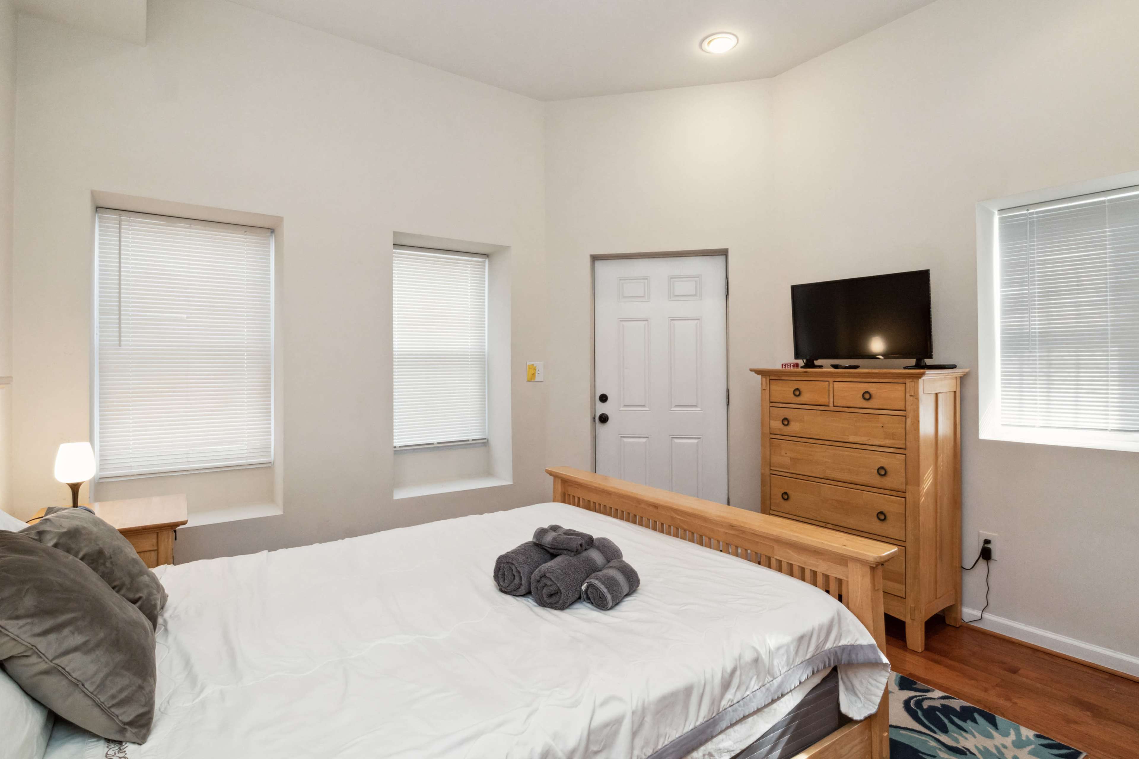A neatly arranged bedroom with a bed, dresser, television, and two windows allowing natural light to enter.