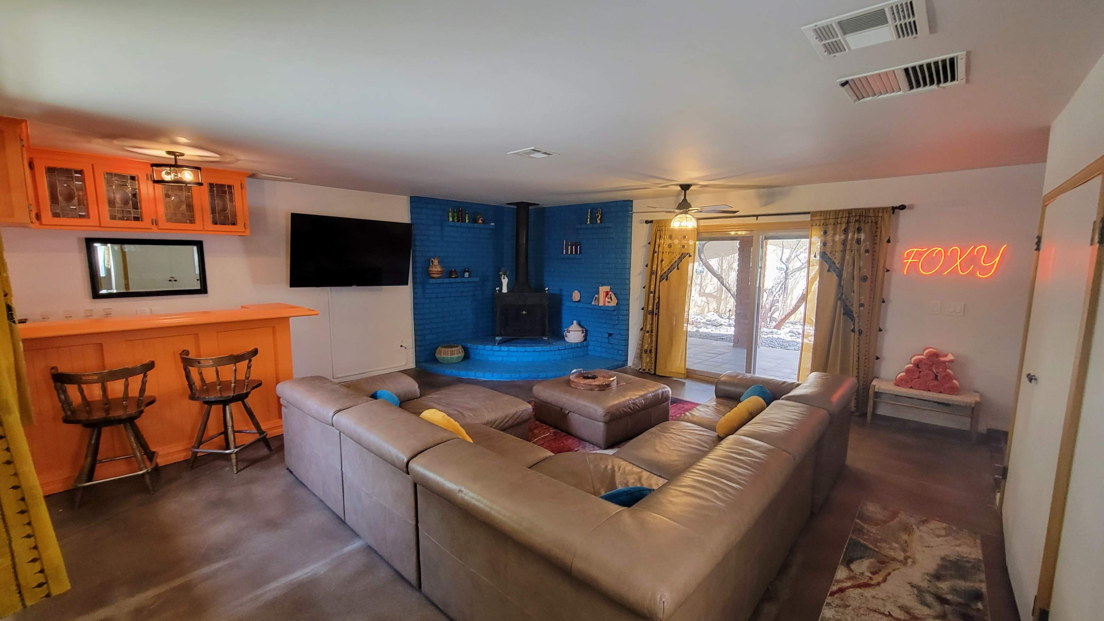 A living room features a large sectional sofa, an orange wet bar, a blue accent wall with a fireplace, and sliding glass doors leading to an outdoor area.