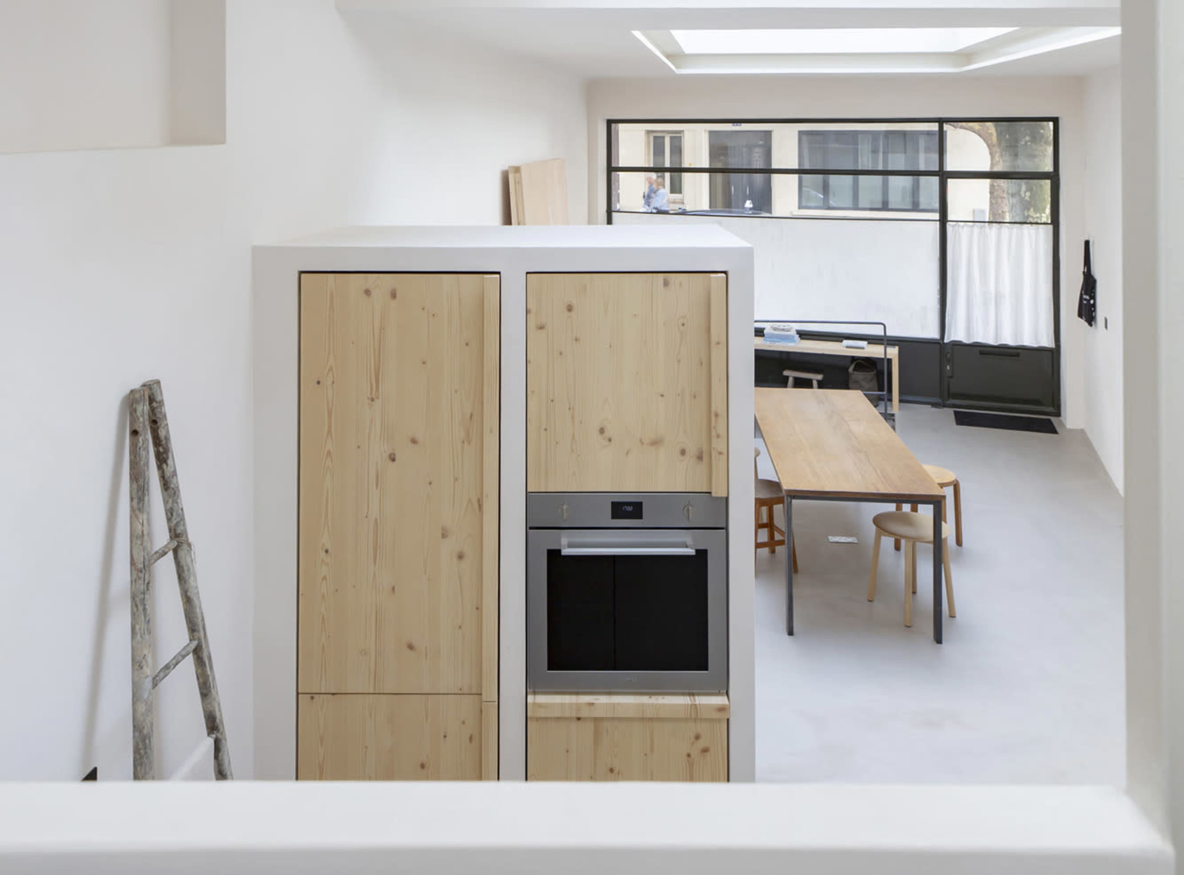 The image shows a modern kitchen with wooden cabinetry, an oven, and a dining area featuring a long table and chairs, illuminated by natural light from a skylight.