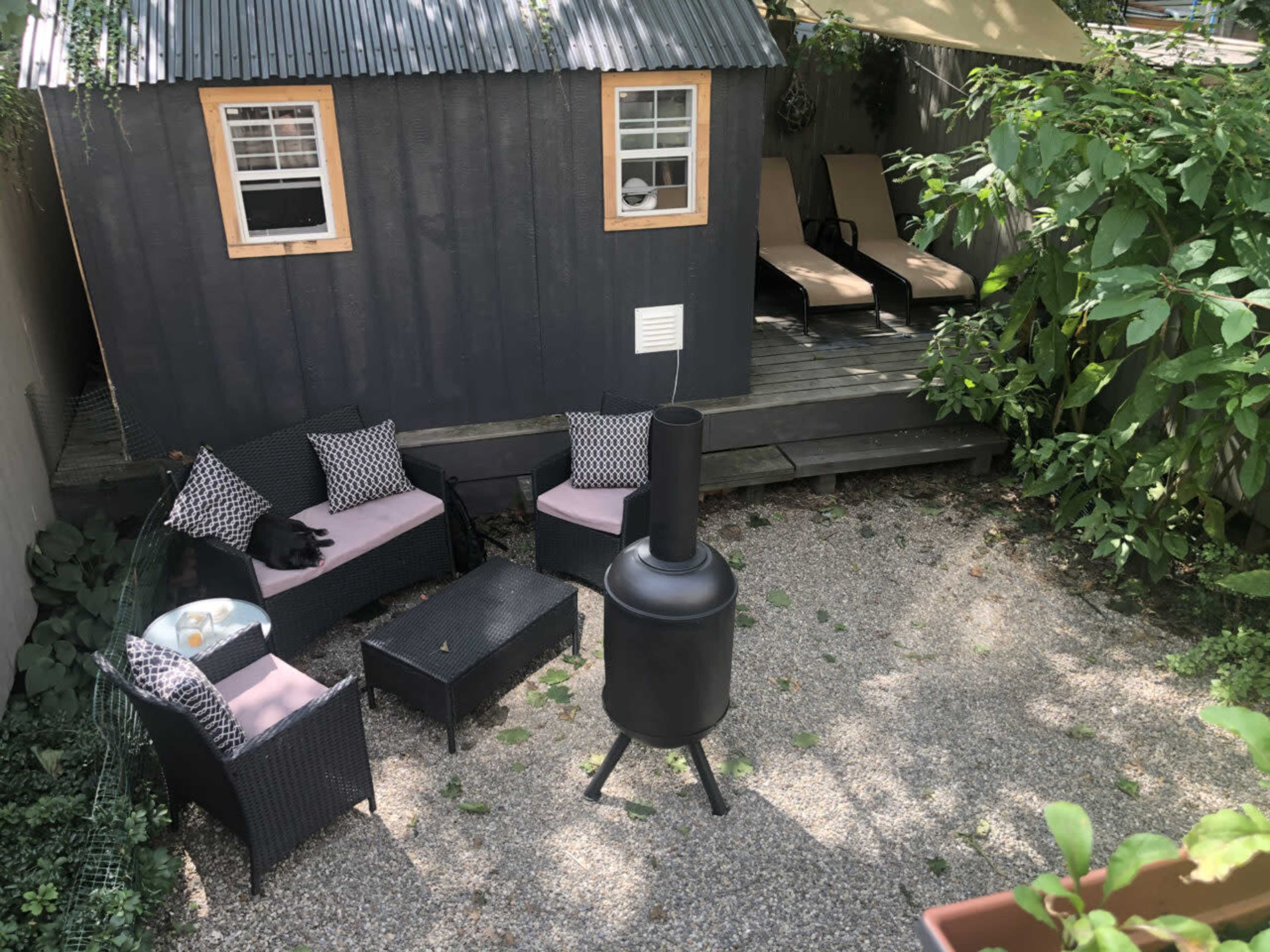 The scene shows a cozy outdoor space featuring a black shed, a gravel patio with seating, and a chimney-style fire pit surrounded by greenery.