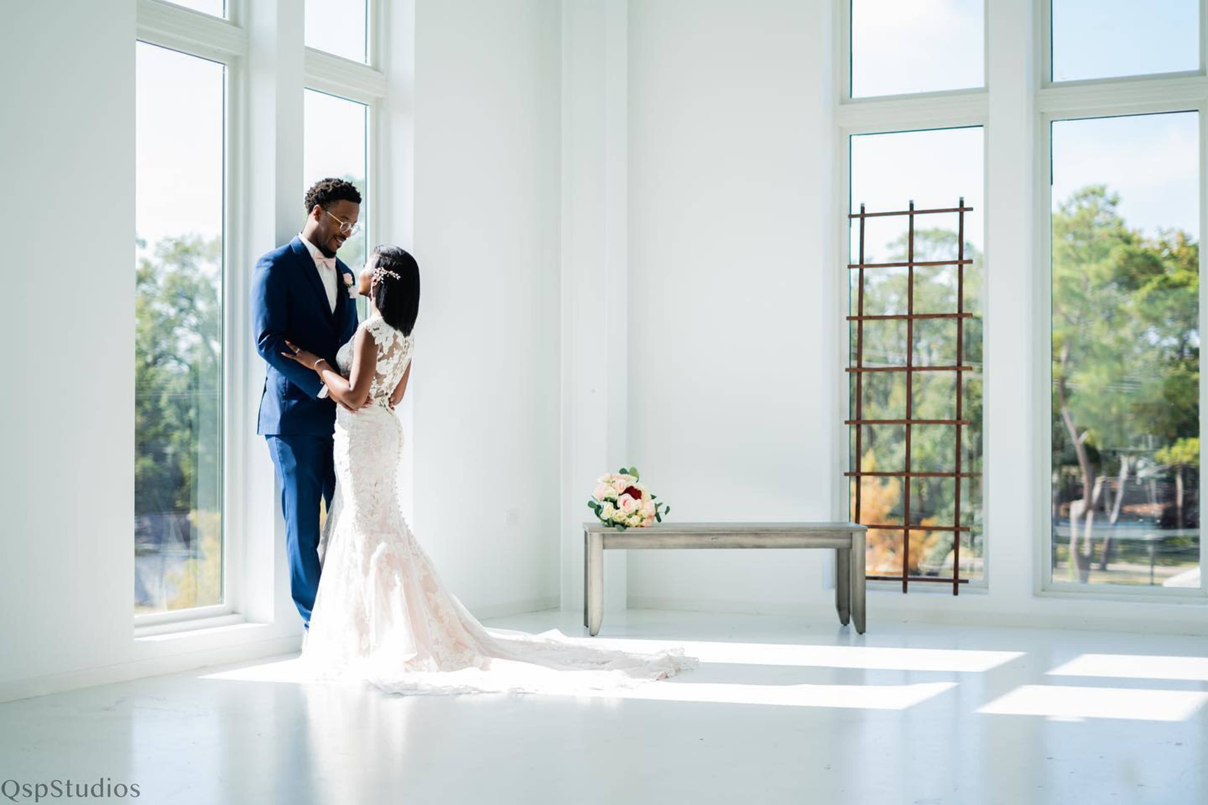 A couple dressed in wedding attire stands together in a bright, modern room with large windows and a simple wooden bench.