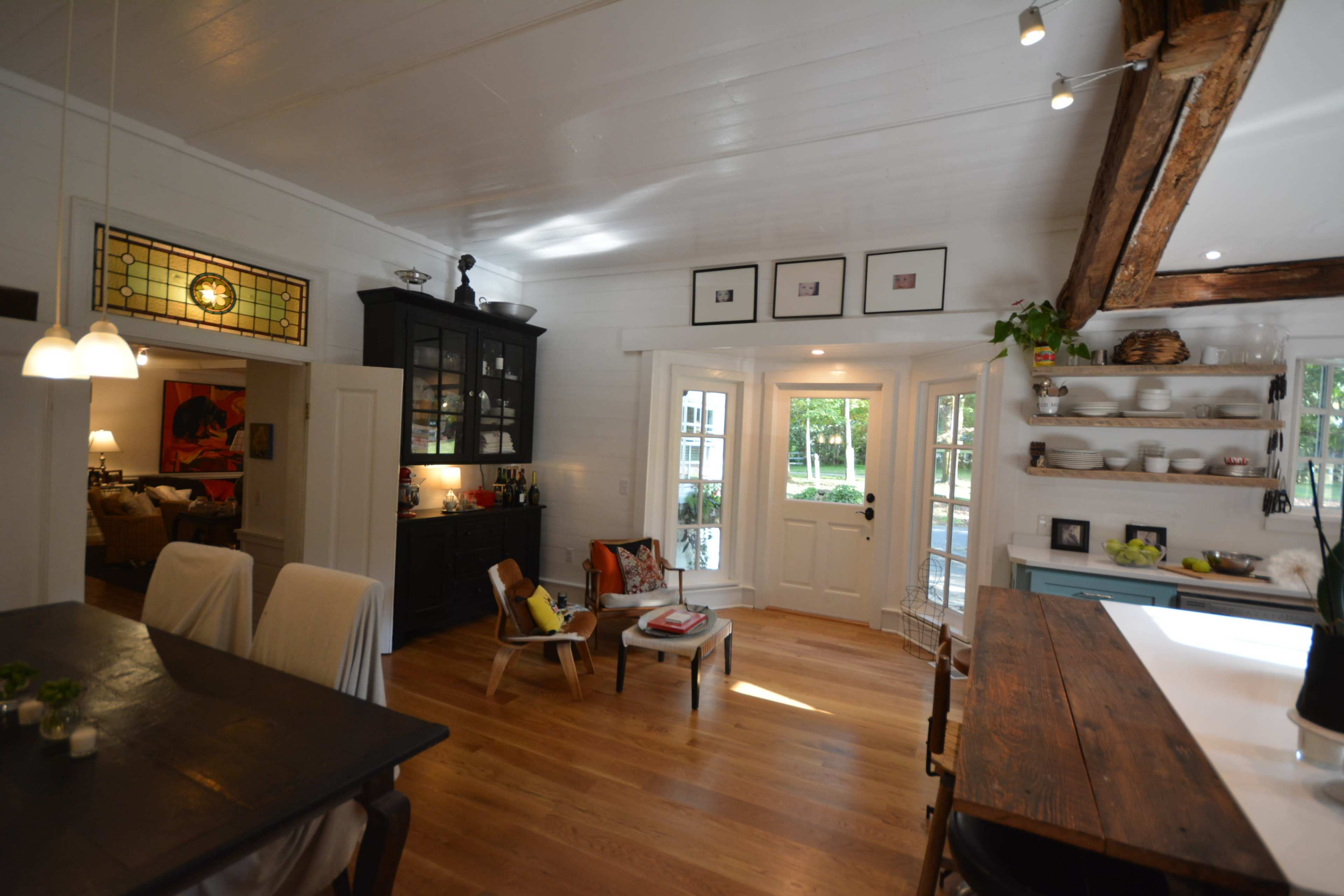 The image shows a bright, open kitchen and dining area with wooden floors, a large window, and a door leading to an outdoor space.