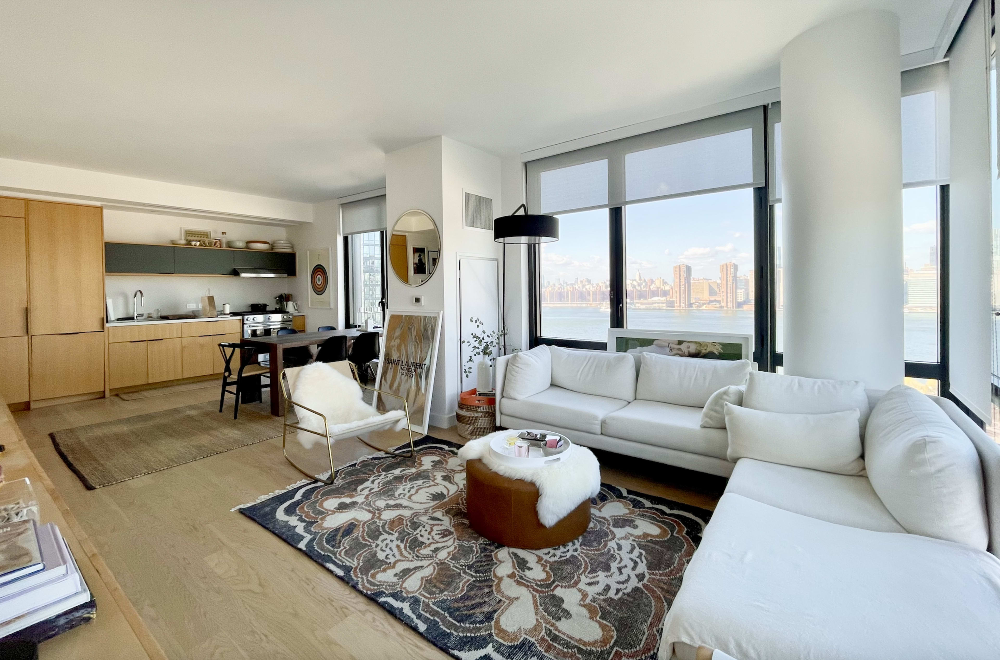 The image shows a modern living room with a large window overlooking a city skyline, featuring a white sectional sofa, a round coffee table, a dining area, and wooden cabinetry.