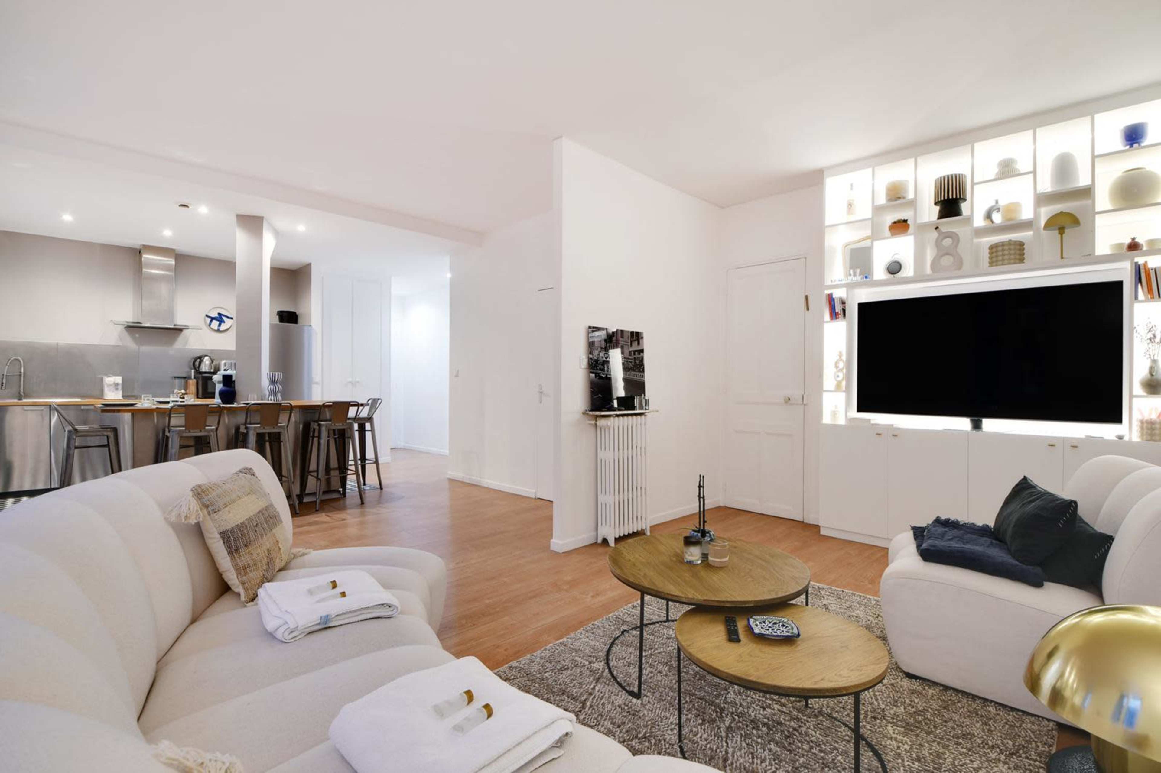 A modern living room features a light-colored sofa, a round coffee table, and a large TV unit, with an open kitchen area visible in the background.