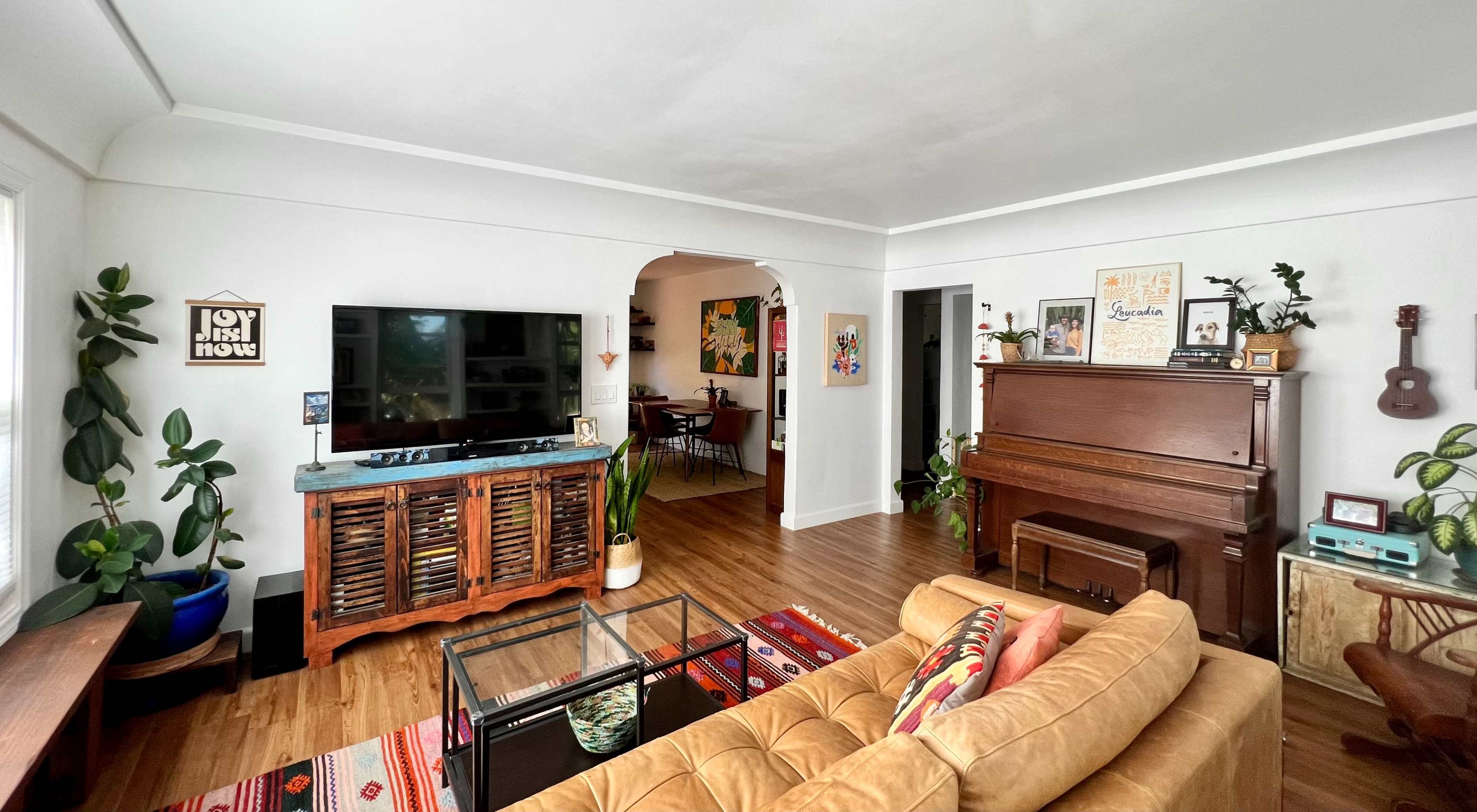 A living room with a sofa, a television on a wooden cabinet, a piano, and potted plants, leading to a dining area in the background.