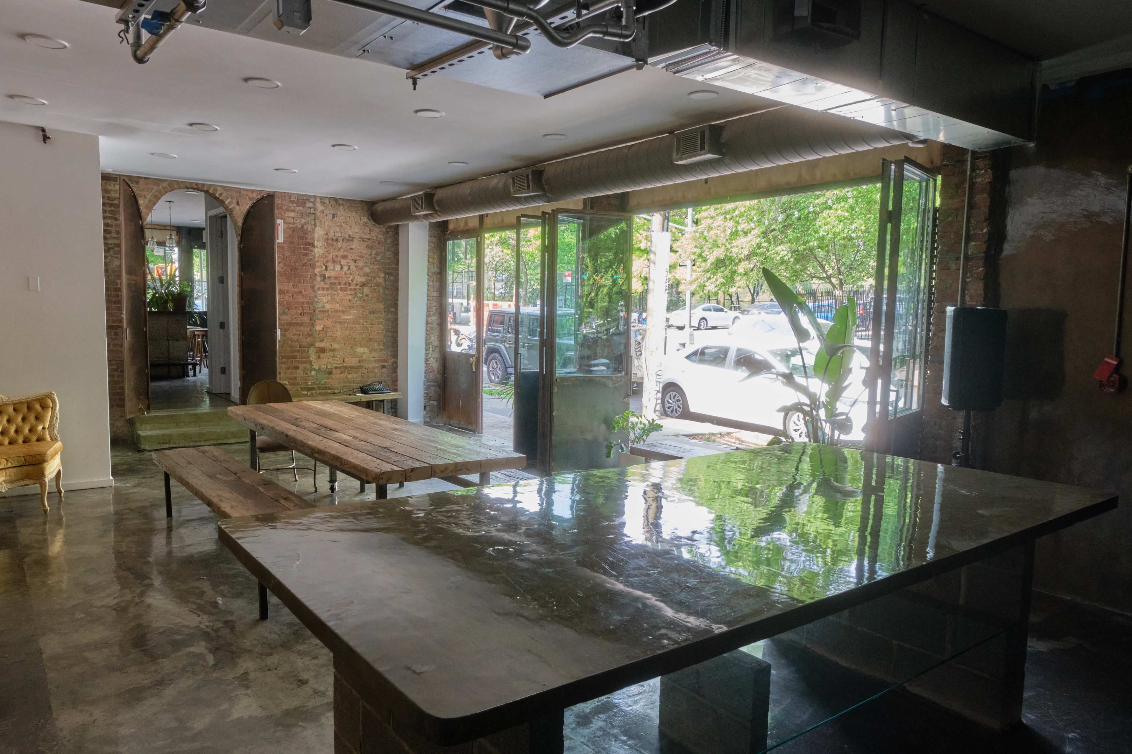 A modern interior space with large glass doors opening to a street view, featuring a central dark table, wooden benches, and potted plants.
