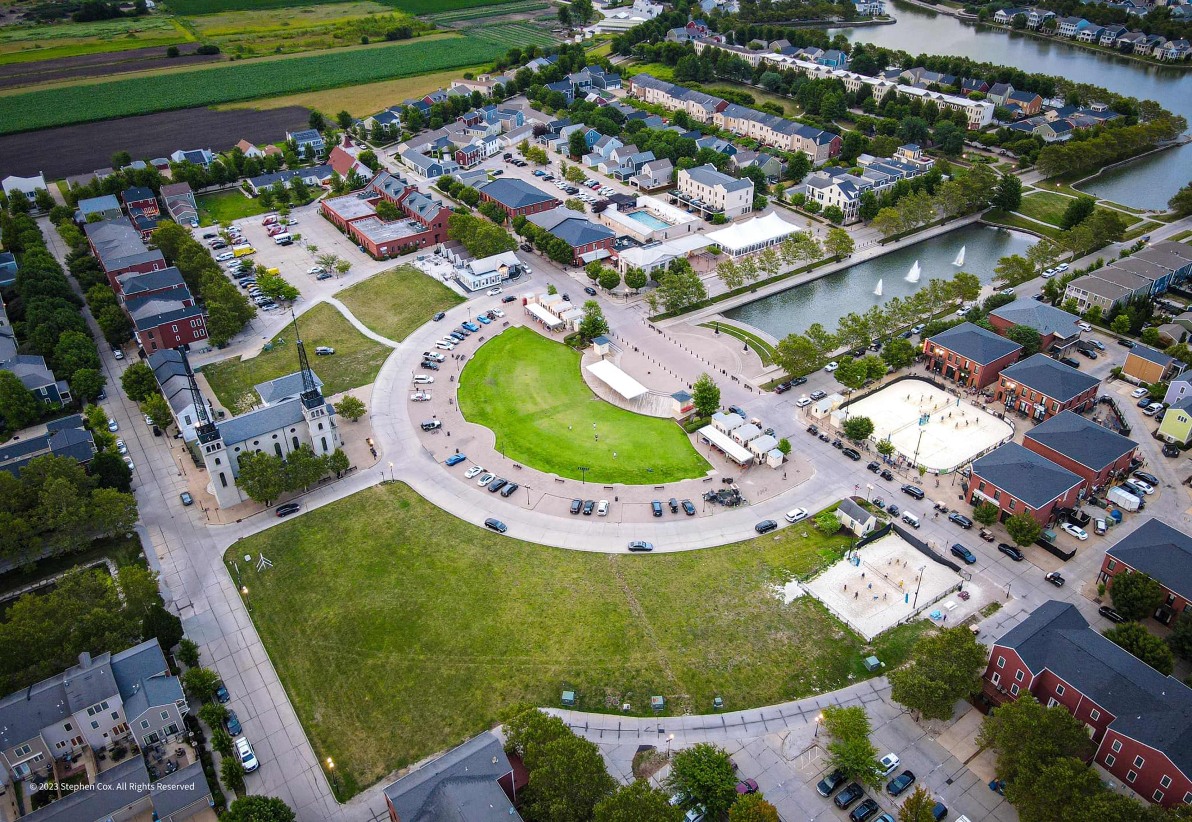 An aerial view shows a residential area featuring a circular green space surrounded by roads, parked cars, and buildings, with a water body nearby.