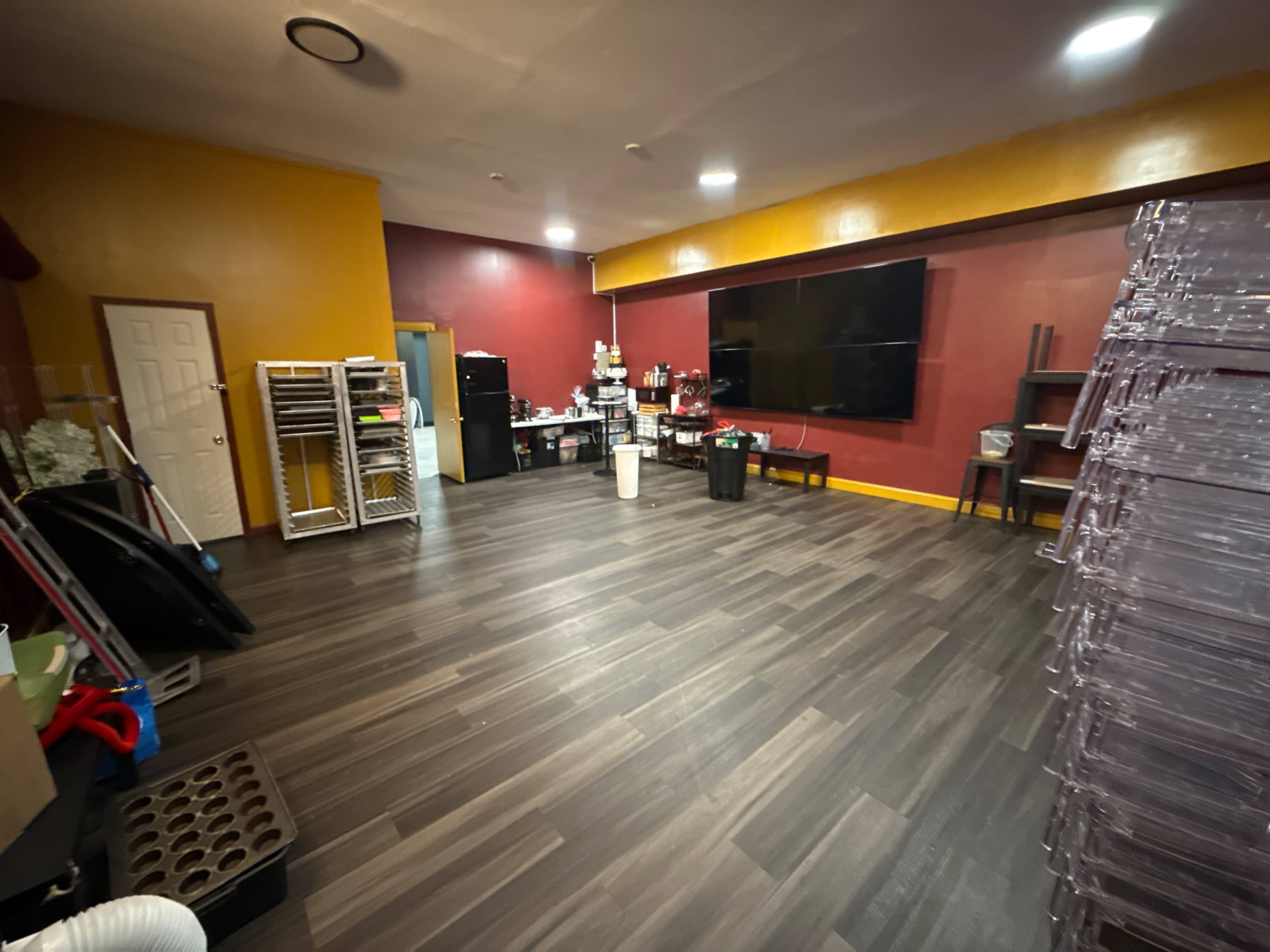 The image shows a spacious room with a mix of yellow and red walls, wooden flooring, and various furniture items including a television, shelving units, and storage containers.
