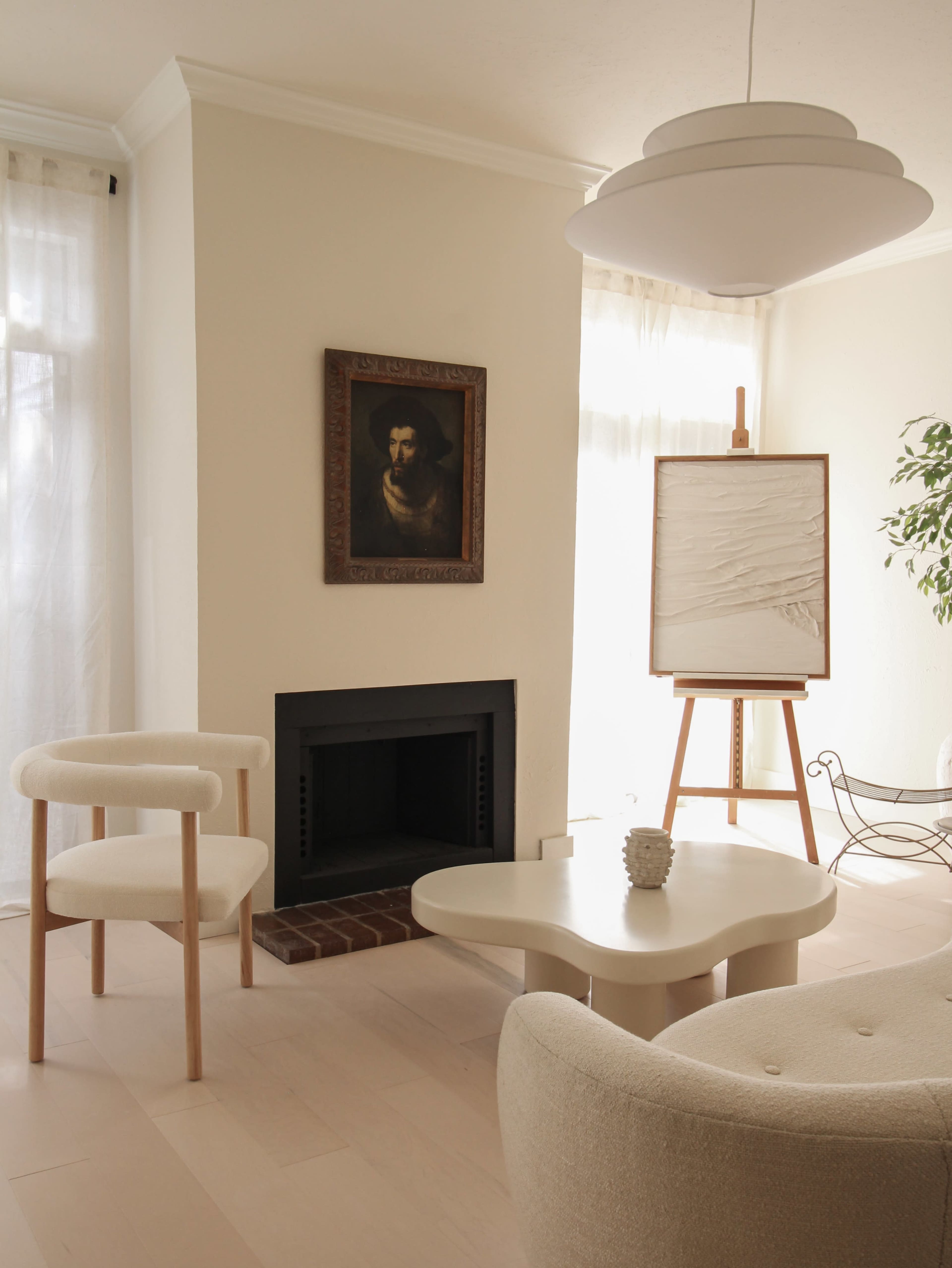 The image shows a minimalist living room featuring a fireplace, a chair, a table, and an easel with a blank canvas.