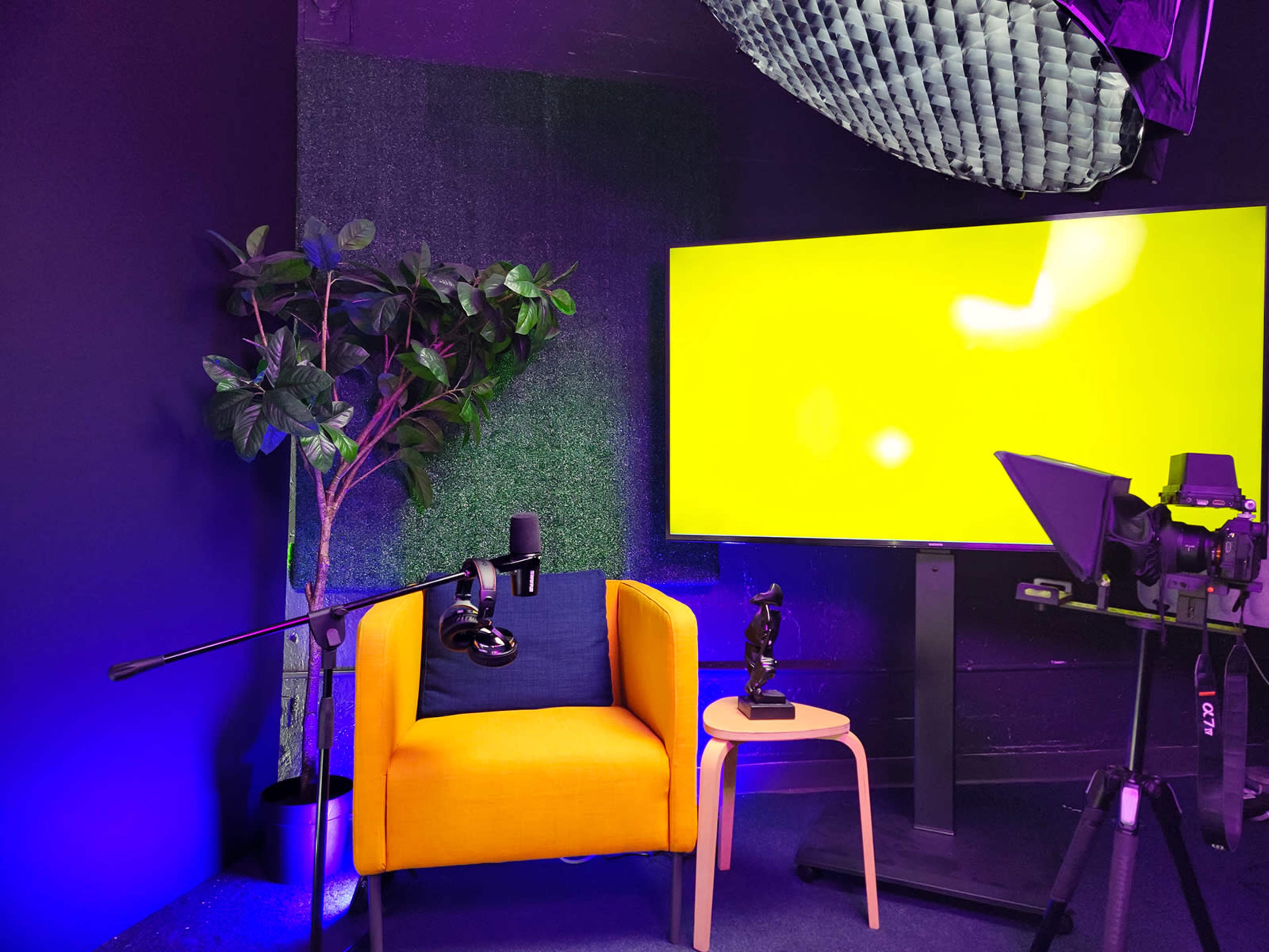 The scene features a brightly colored yellow chair positioned next to a large yellow screen, with a microphone, camera, and a small plant in the background.