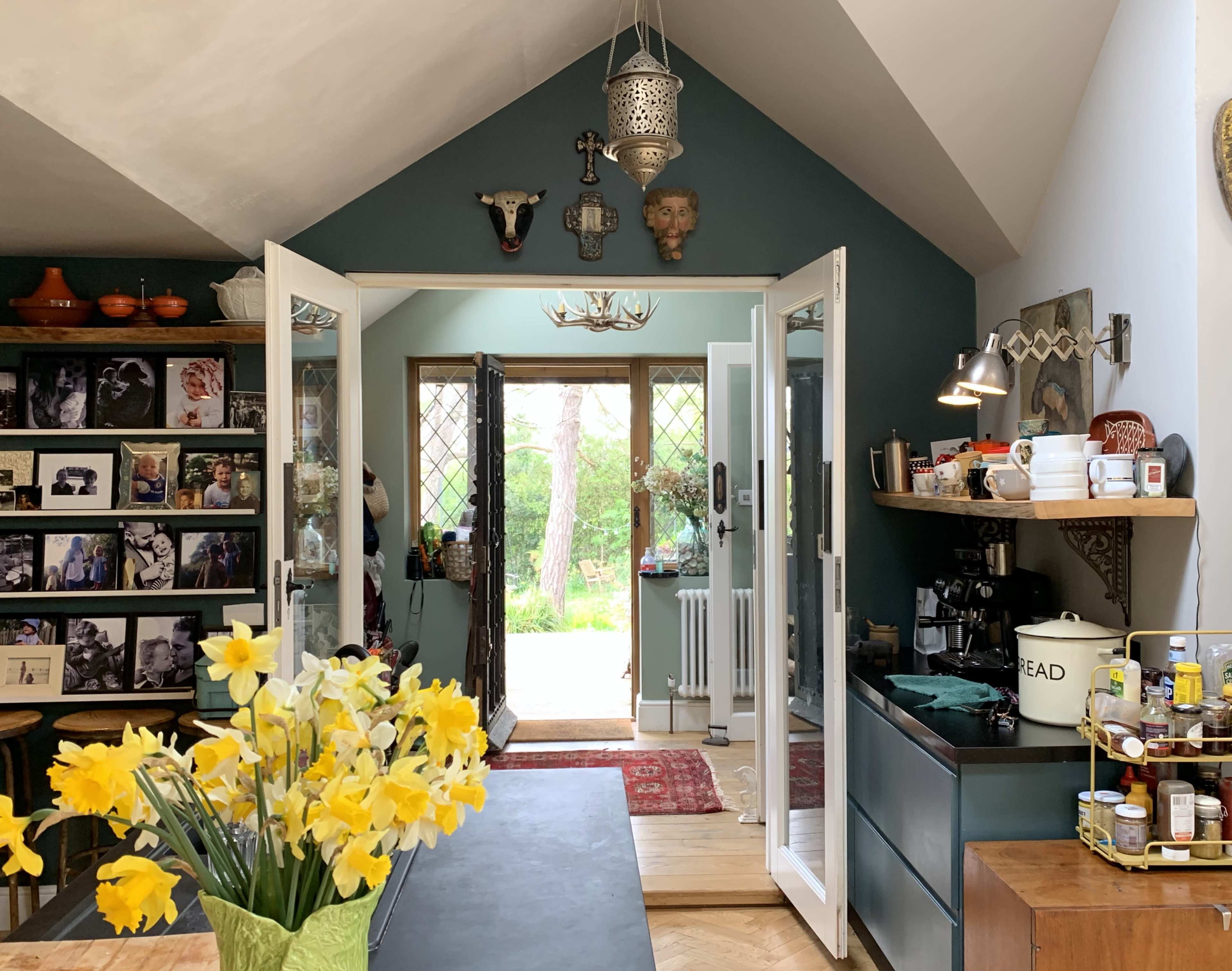 A cozy kitchen features open double doors leading to a garden, with a table adorned with yellow flowers and walls decorated with various photographs and quirky decor.