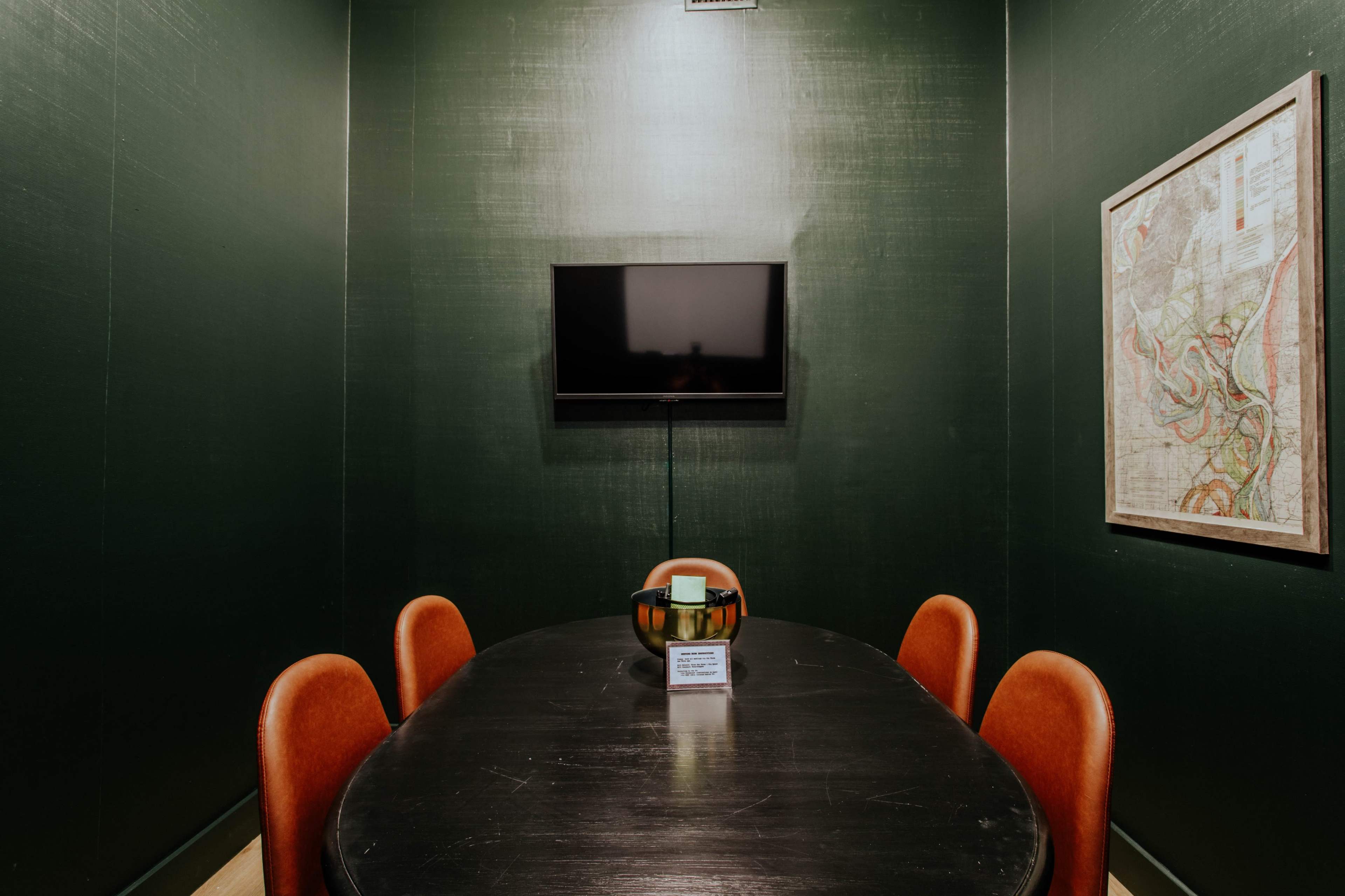 A small conference room with a dark green wall, a black table surrounded by orange chairs, and a mounted television.